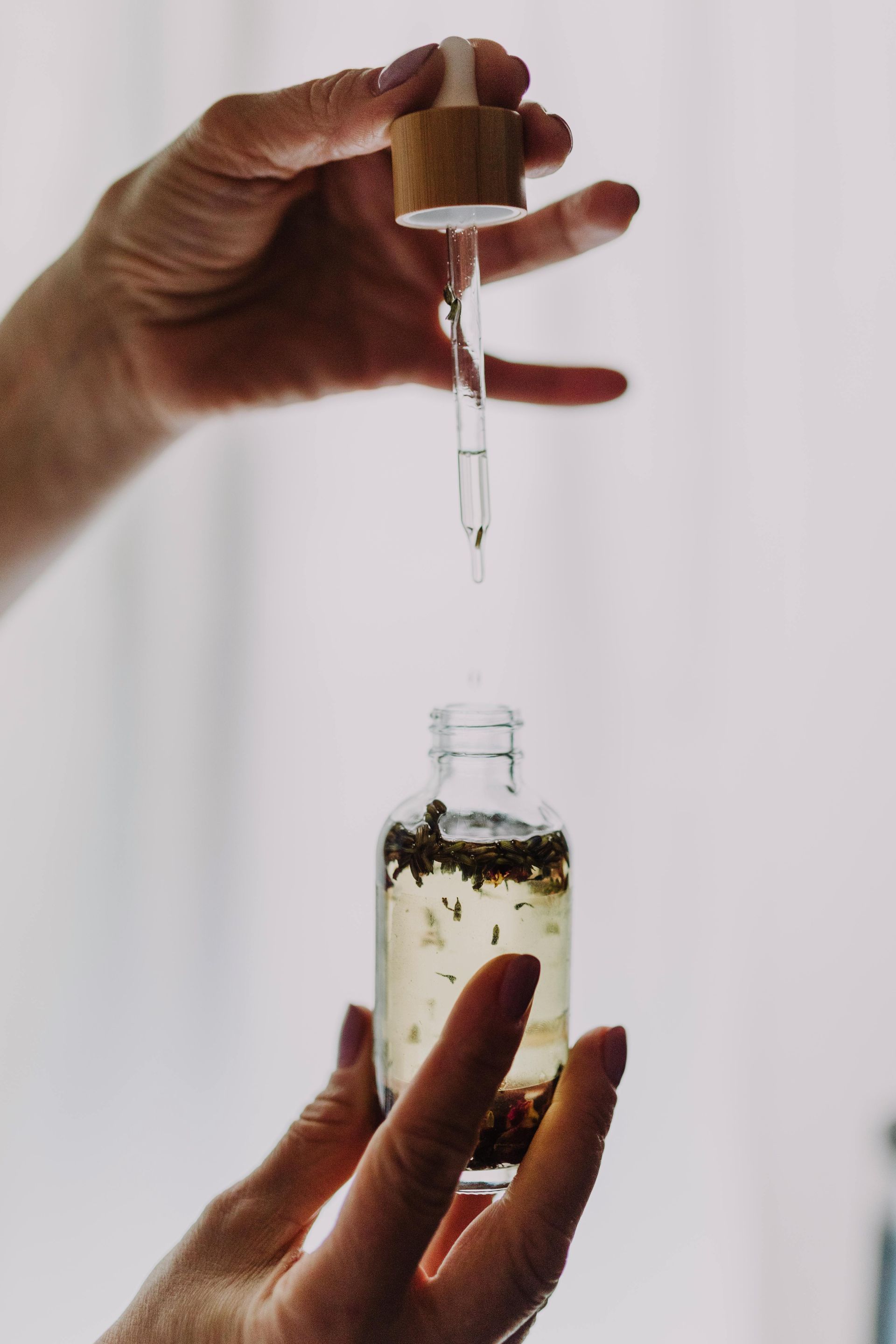 Hands holding a dropper with liquid dripping into a small glass bottle filled with botanicals.