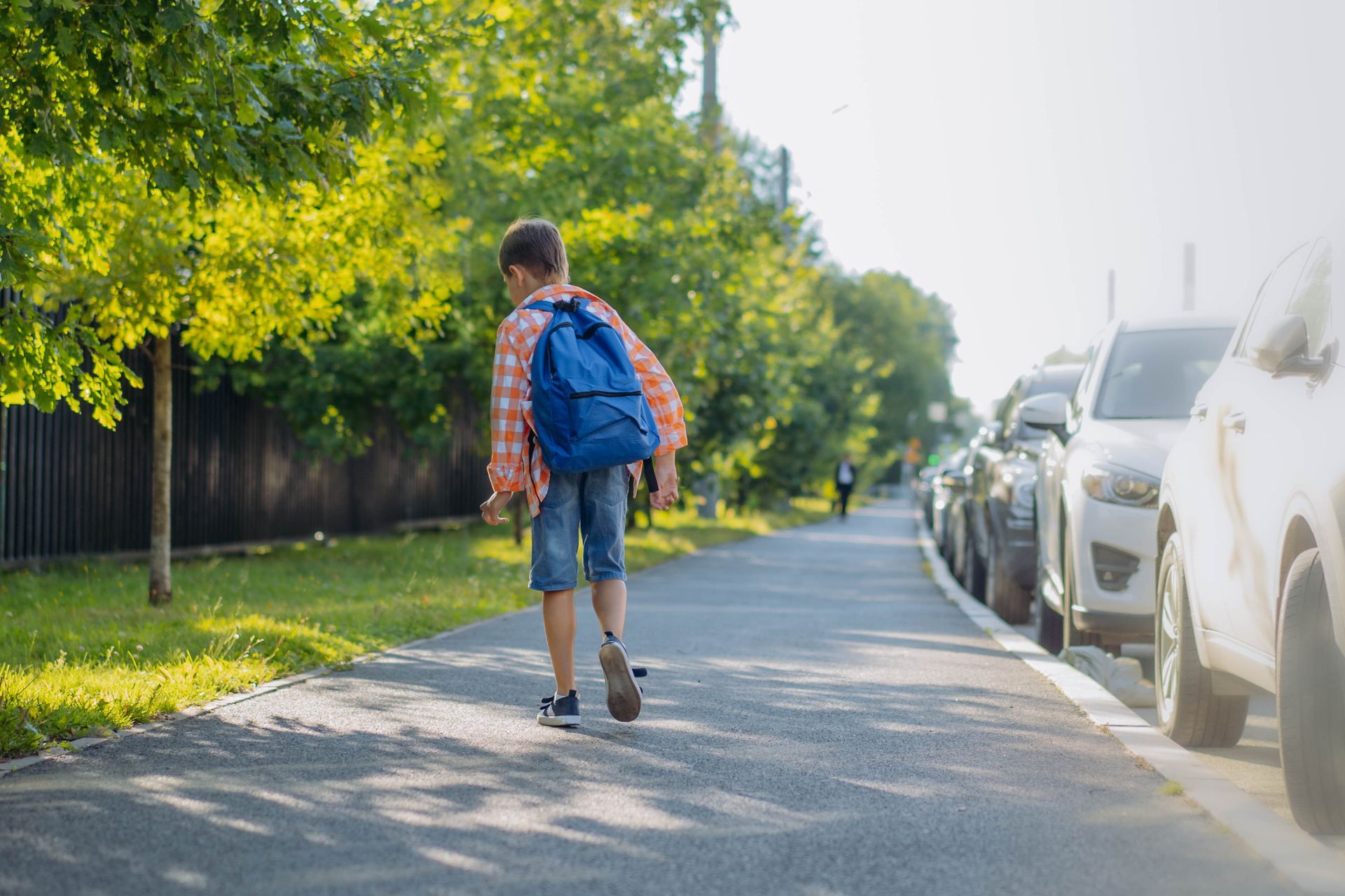Boy walking on sidewalk, blue backpack, towards cars parked on street. Sunlight.