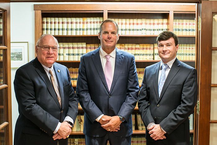 A park path lined with trees, benches, and lampposts. Sunlight filters through the green and brown foliage.