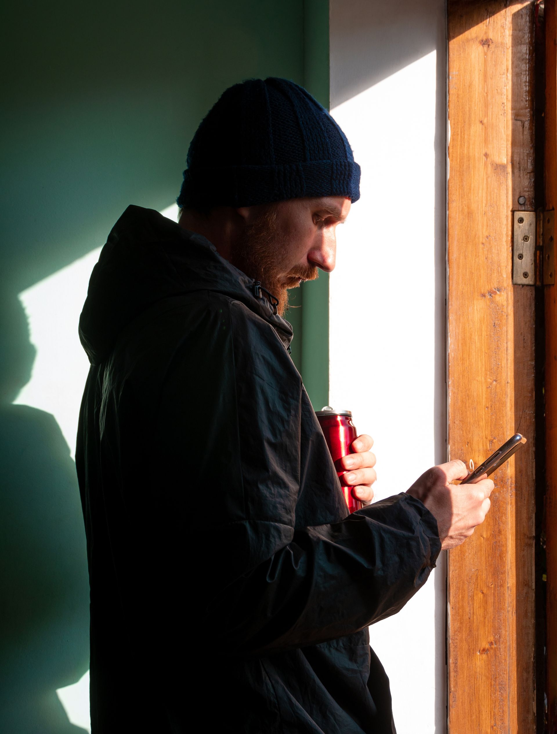 Man in dark jacket and beanie, holding a can and phone, near a doorway, in sunlight.