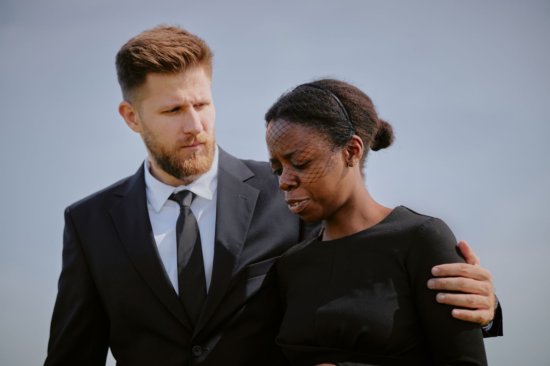 Man in black suit comforts a woman in black dress, both looking somber, outdoors.