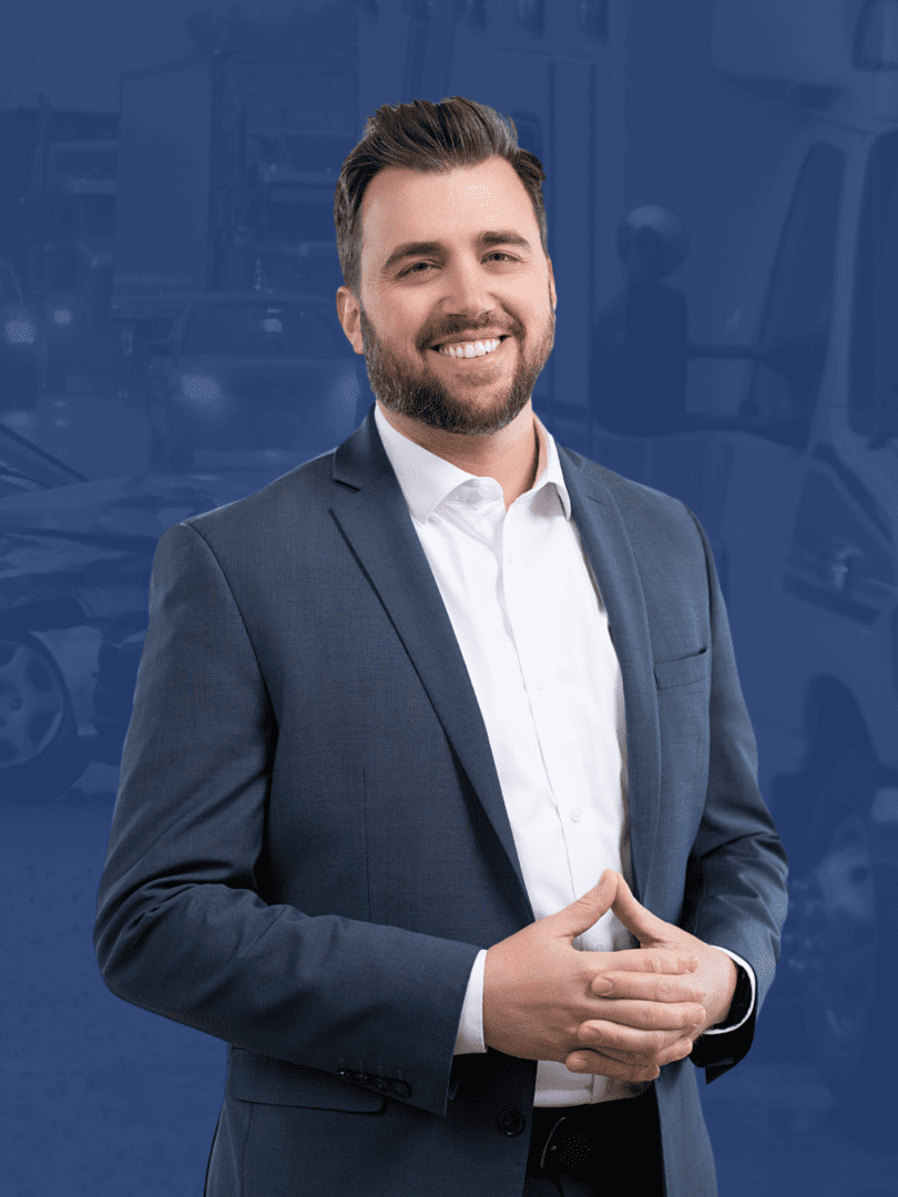 Man in a blue suit smiling, hands clasped. Blurred blue background with vehicles.