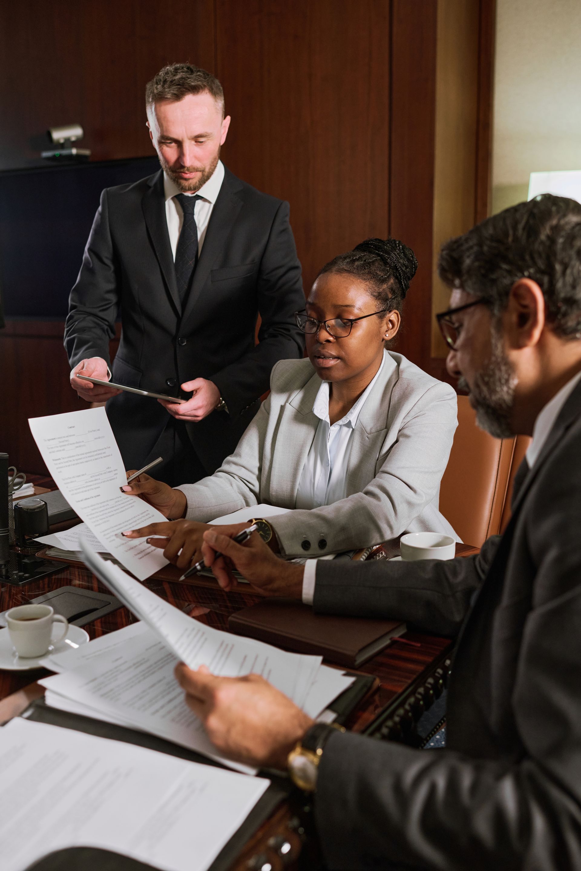 Three people in suits reviewing documents at a table. A man stands, and two others sit.
