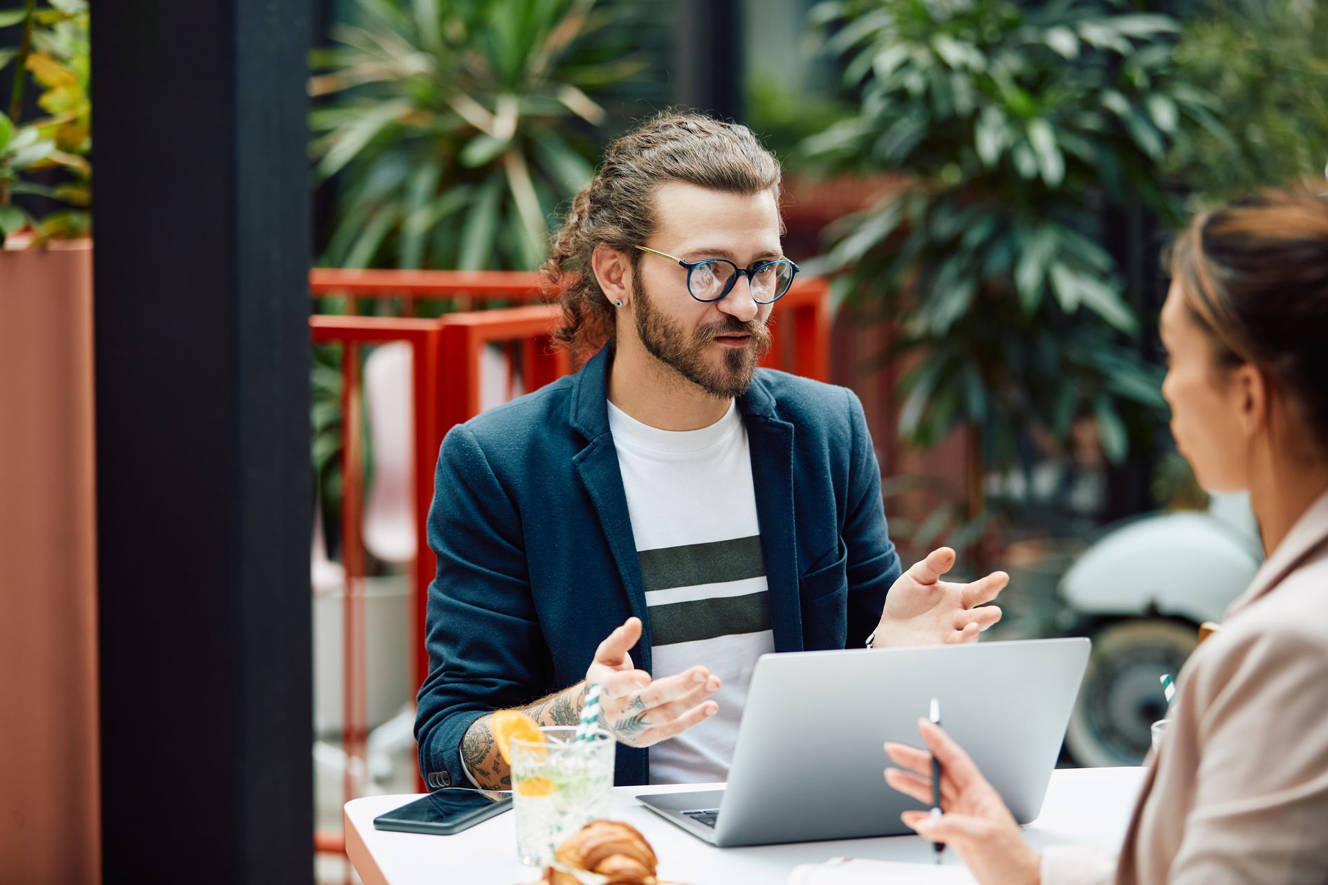 A person with a beard and glasses sits at an outdoor cafe table with a laptop, gesturing while talking to a colleague.