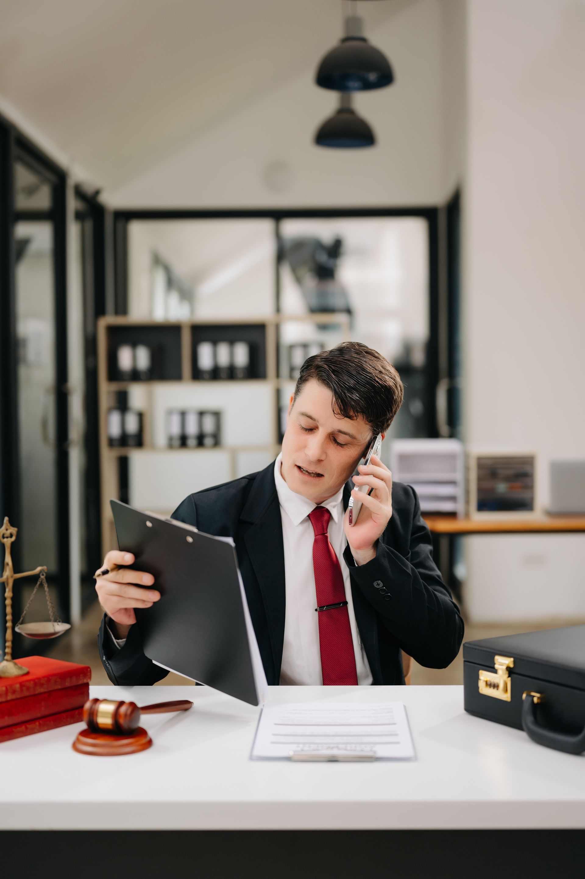 Lawyer at desk holding paperwork, speaking on phone; scales of justice, briefcase visible.
