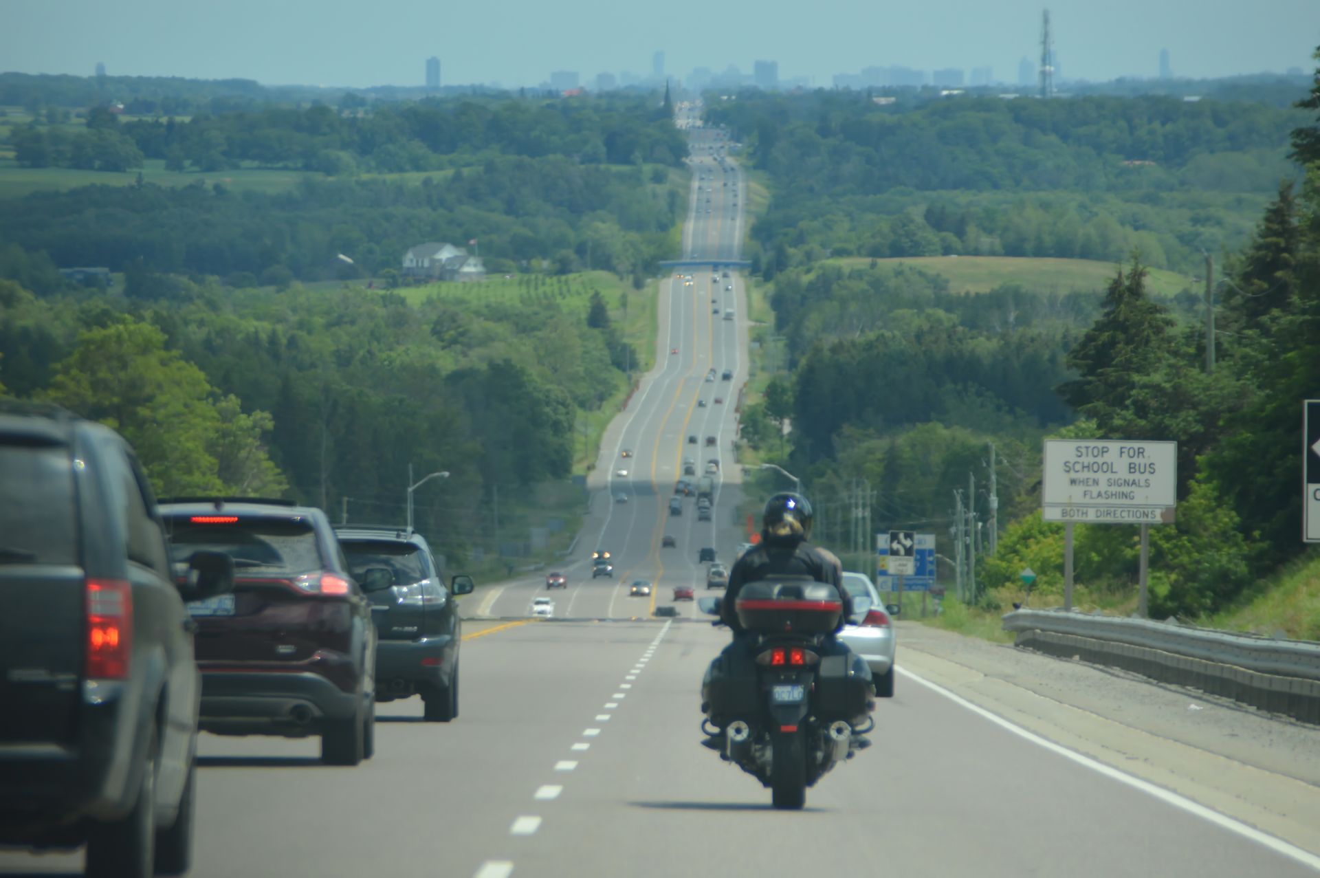 A motorcyclist rides on a highway heading toward a distant cityscape, with cars in adjacent lanes and green hills beyond.