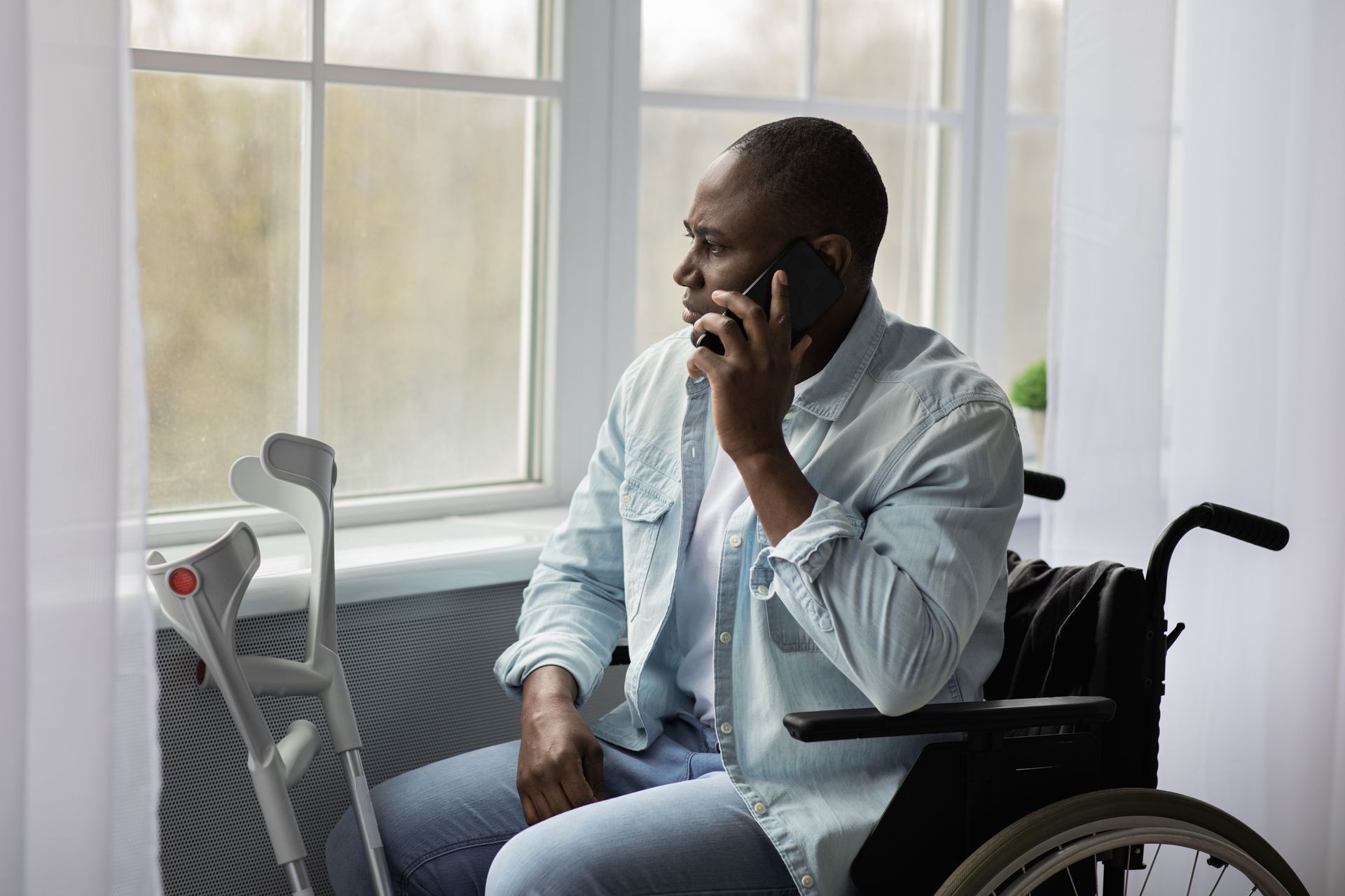 Man in wheelchair talking on phone by window, crutches nearby.
