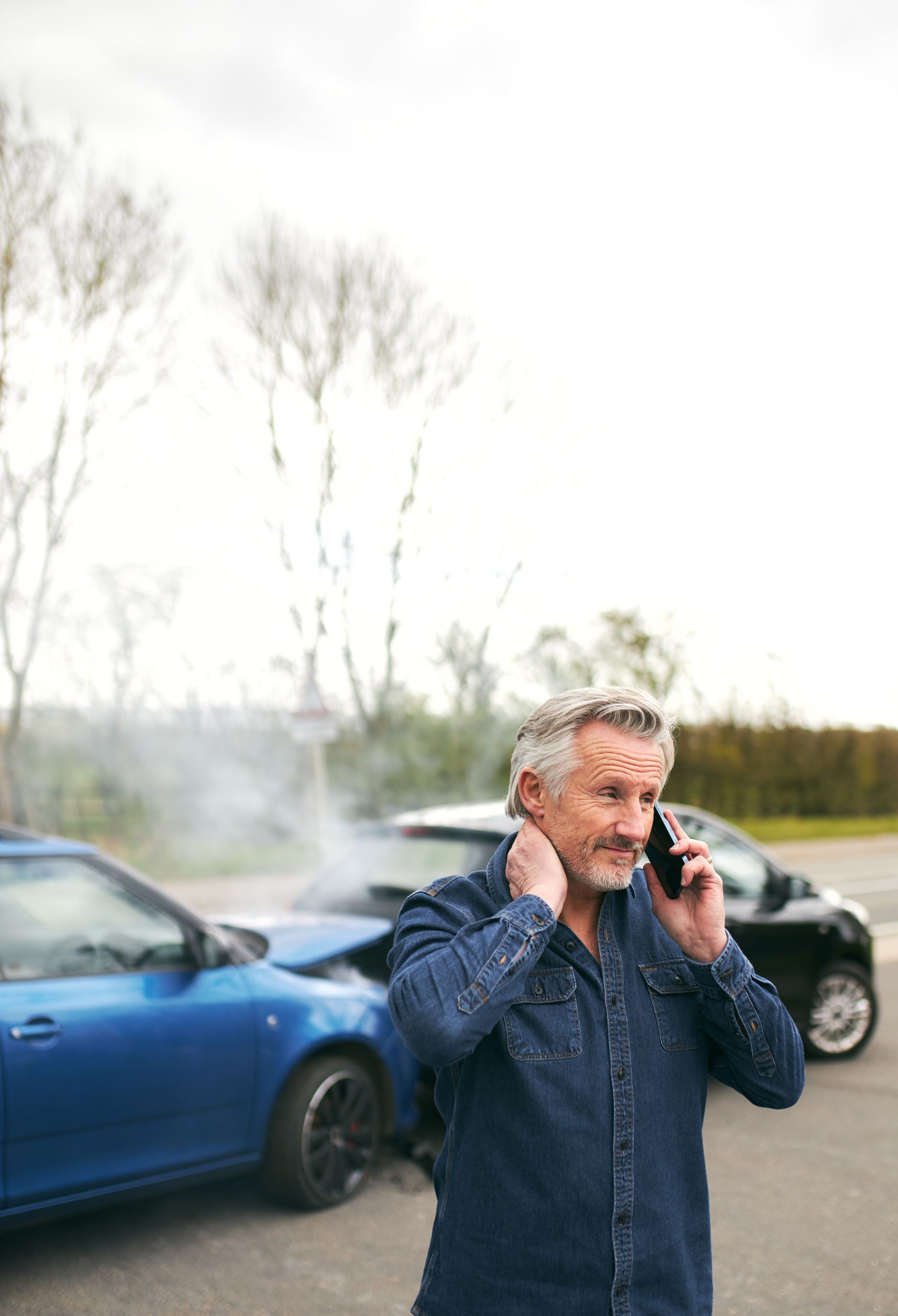 A person speaks on a phone while standing in front of two cars involved in a collision, with smoke rising from the vehicles.