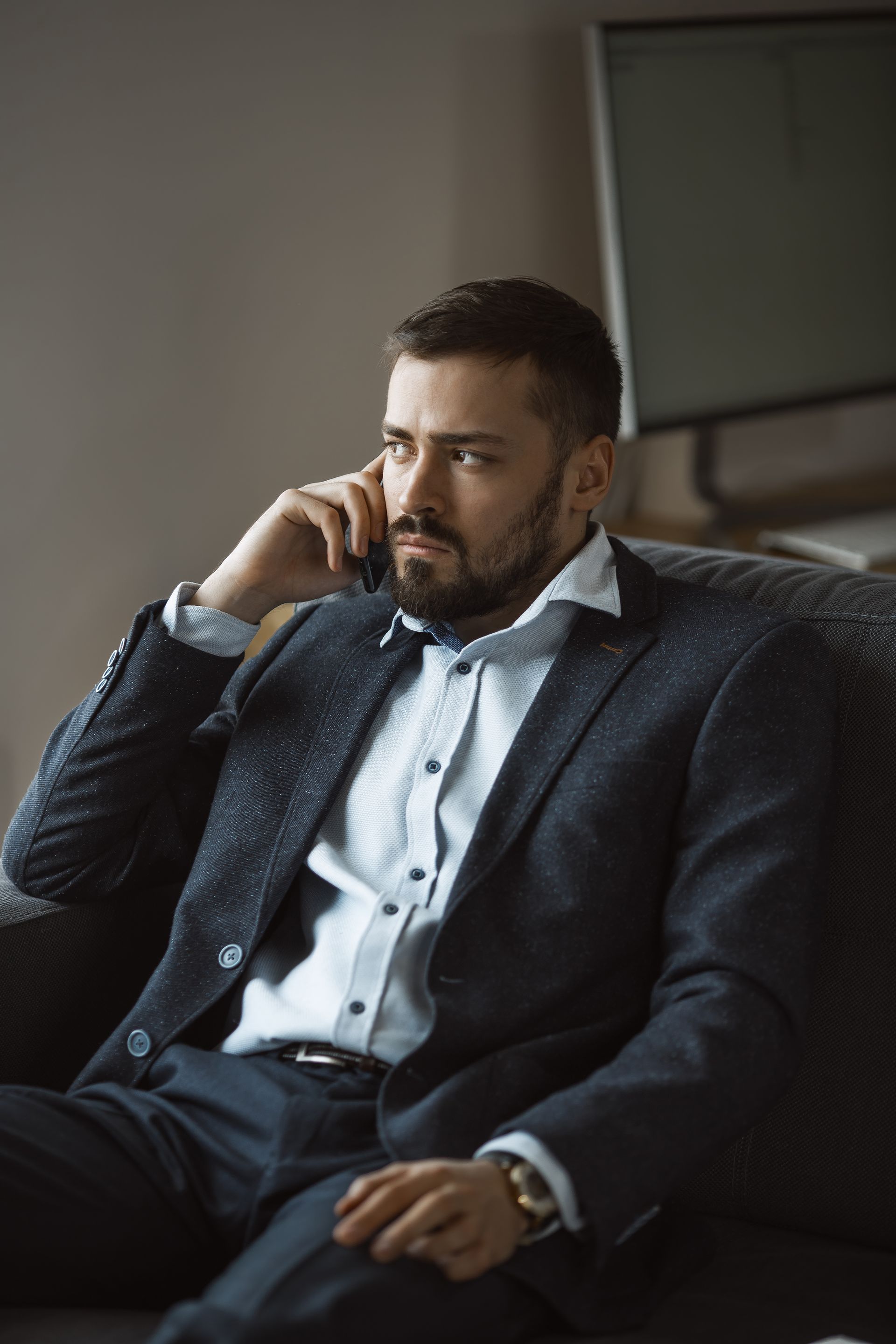A bearded man in a suit and white shirt sits in an office, talking on a smartphone with a serious expression.