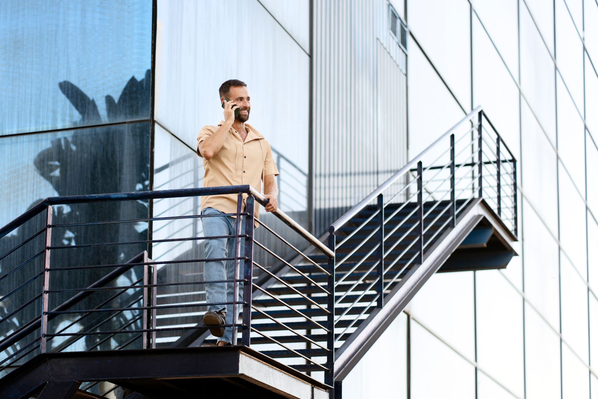 A person in a beige shirt and jeans stands on an outdoor metal staircase, talking on a smartphone against a glass wall.