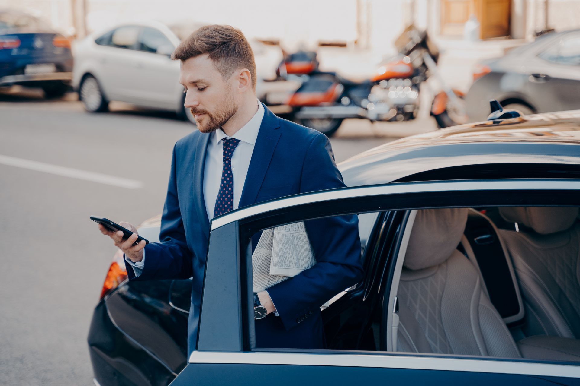 Man in blue suit, checking phone, getting out of black car on street.