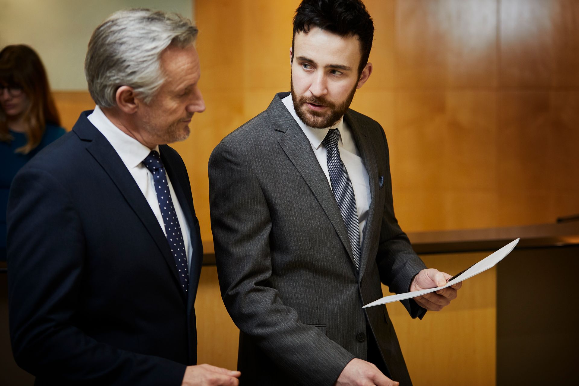 Two men in suits, discussing paperwork in an office setting.