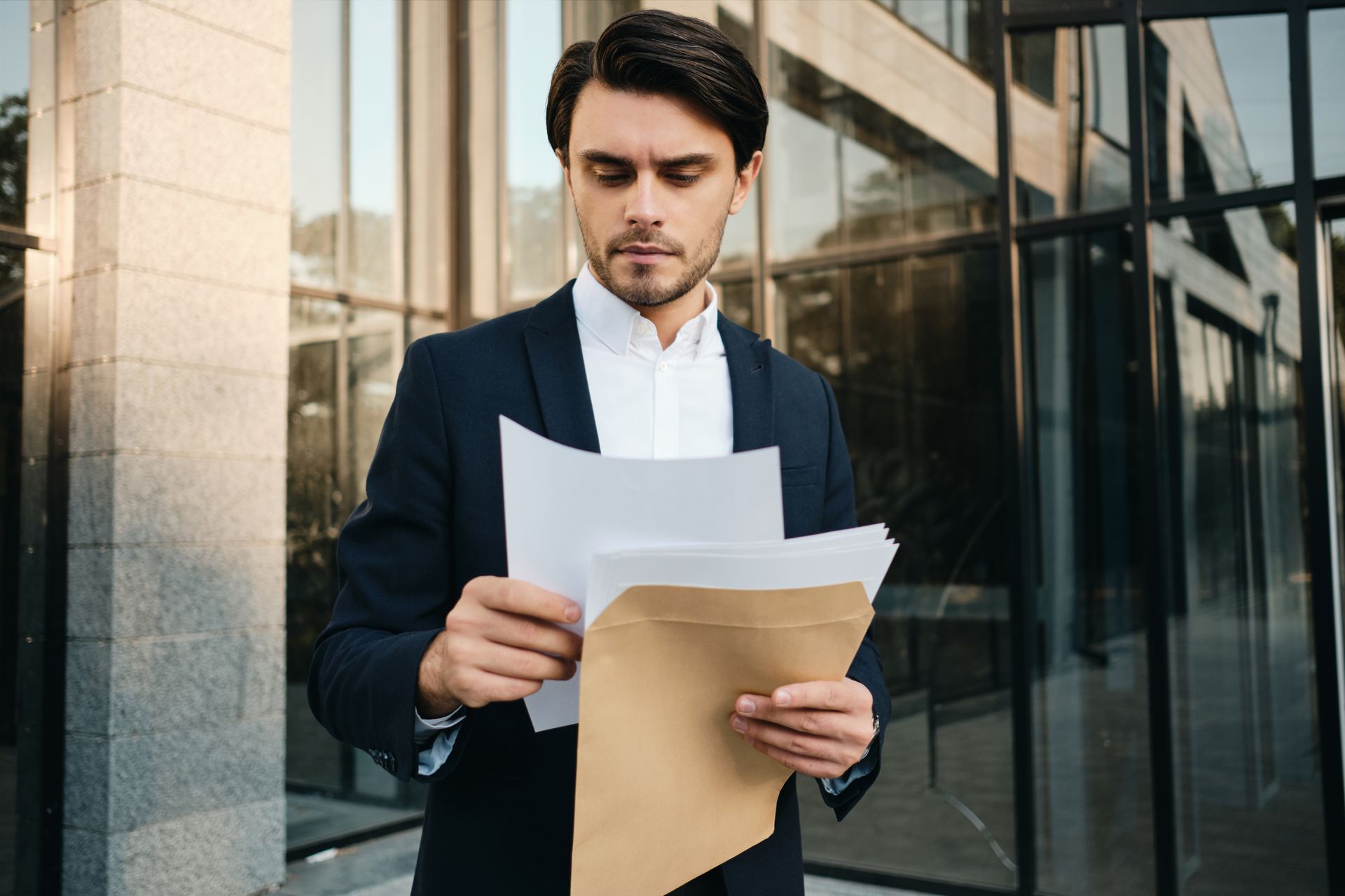 A person in a dark suit and white shirt standing outdoors while reading a document from a tan envelope.