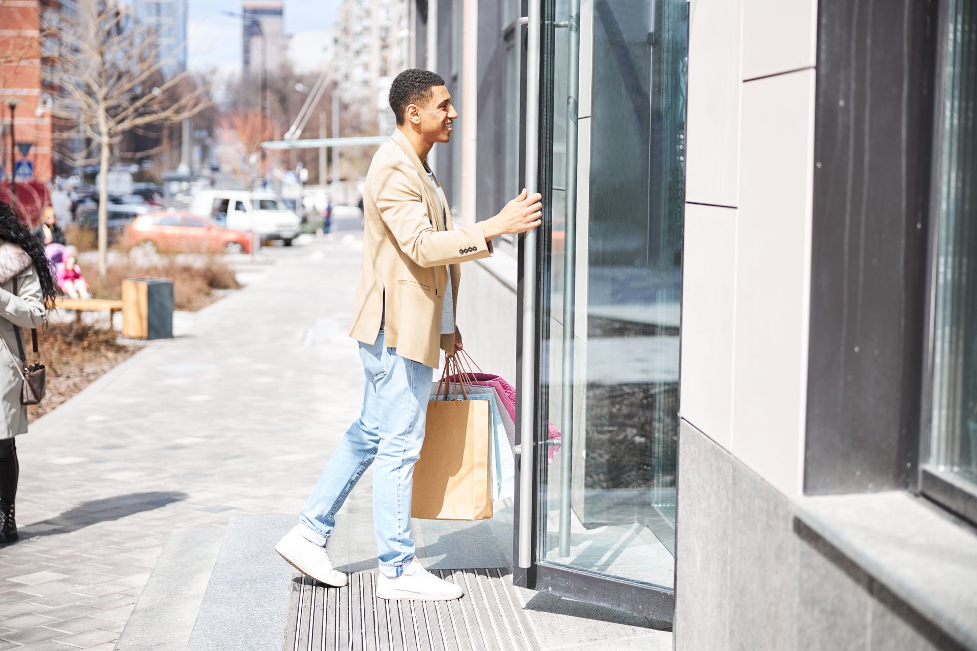 A person in a beige jacket and light blue jeans holds shopping bags while opening a glass door on a city street.