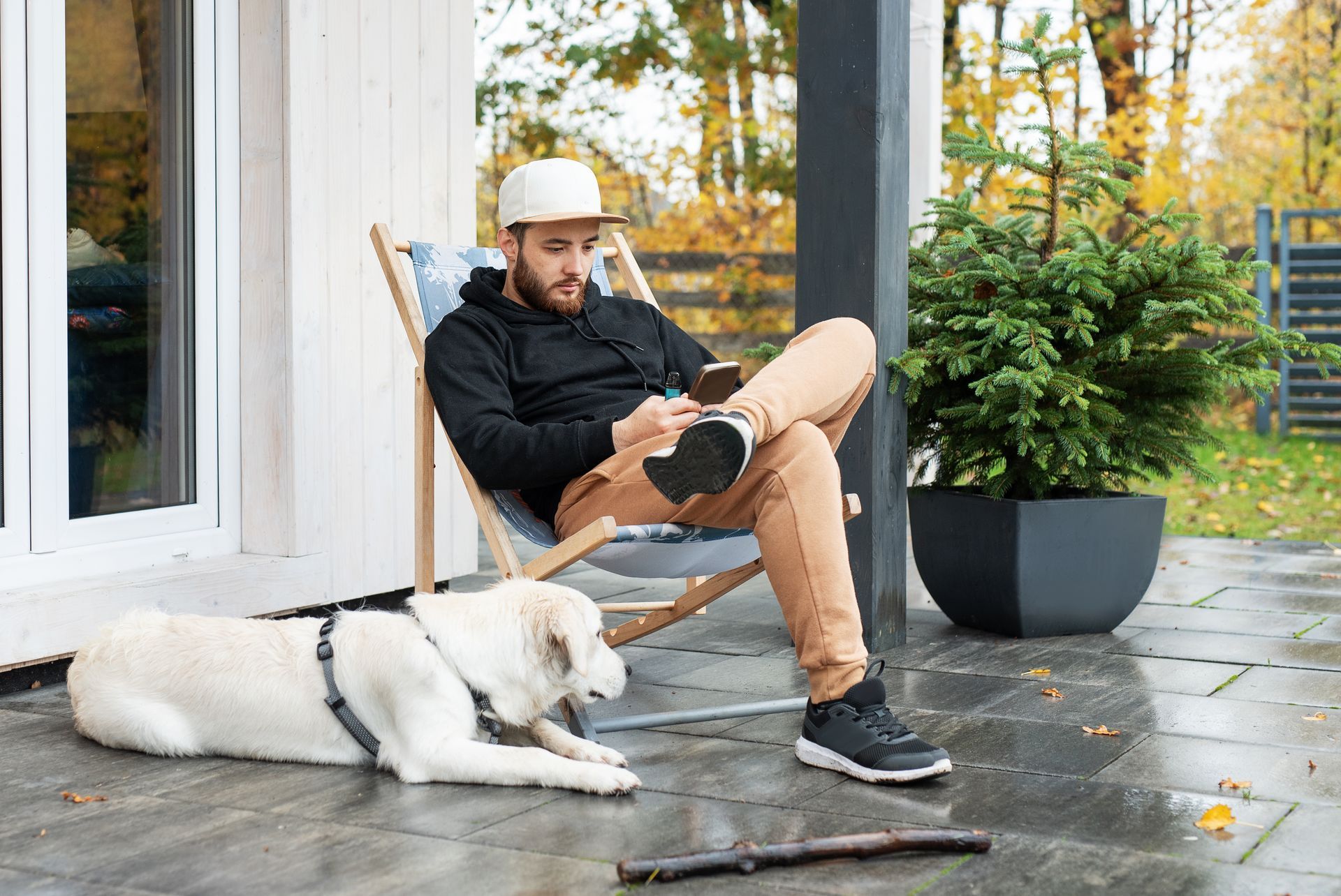 A person in a black hoodie and cap sits in a deck chair using a phone, with a white dog resting on the patio nearby.
