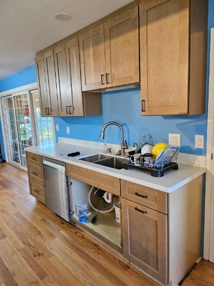 Bright kitchen galley with wood cabinets, blue backsplash, white counters, sink, and dishwasher