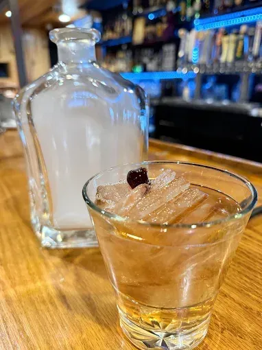 Cocktail on a bar, with smoky decanter behind it. Brown liquid, ice, garnish. Bar in background.