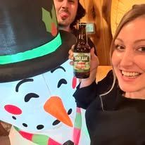Person with beer and inflatable snowman smiles next to a woman; setting is indoors.