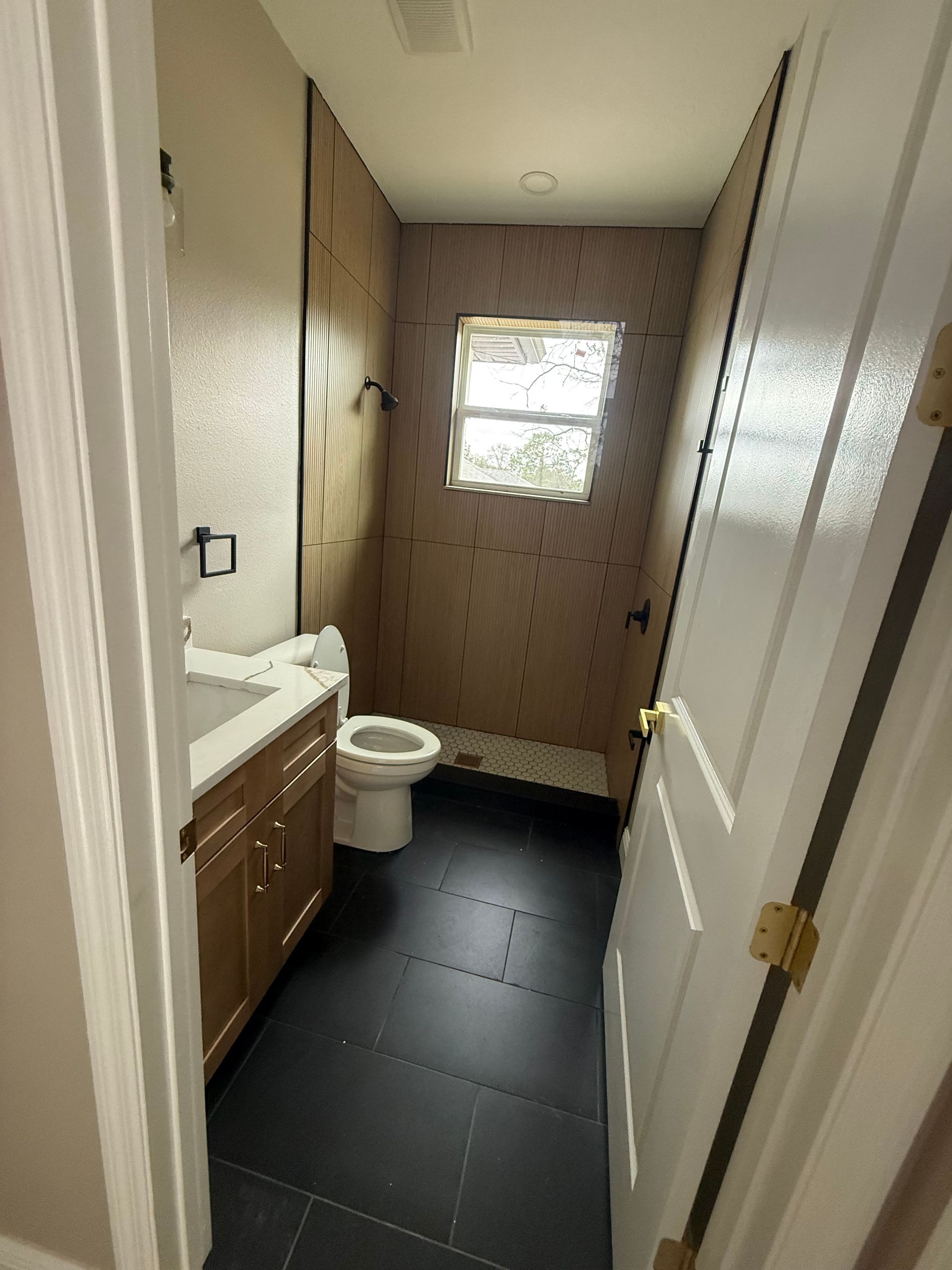 Small bathroom with white toilet, dark tile floor, wood vanity, and a window at the far end.