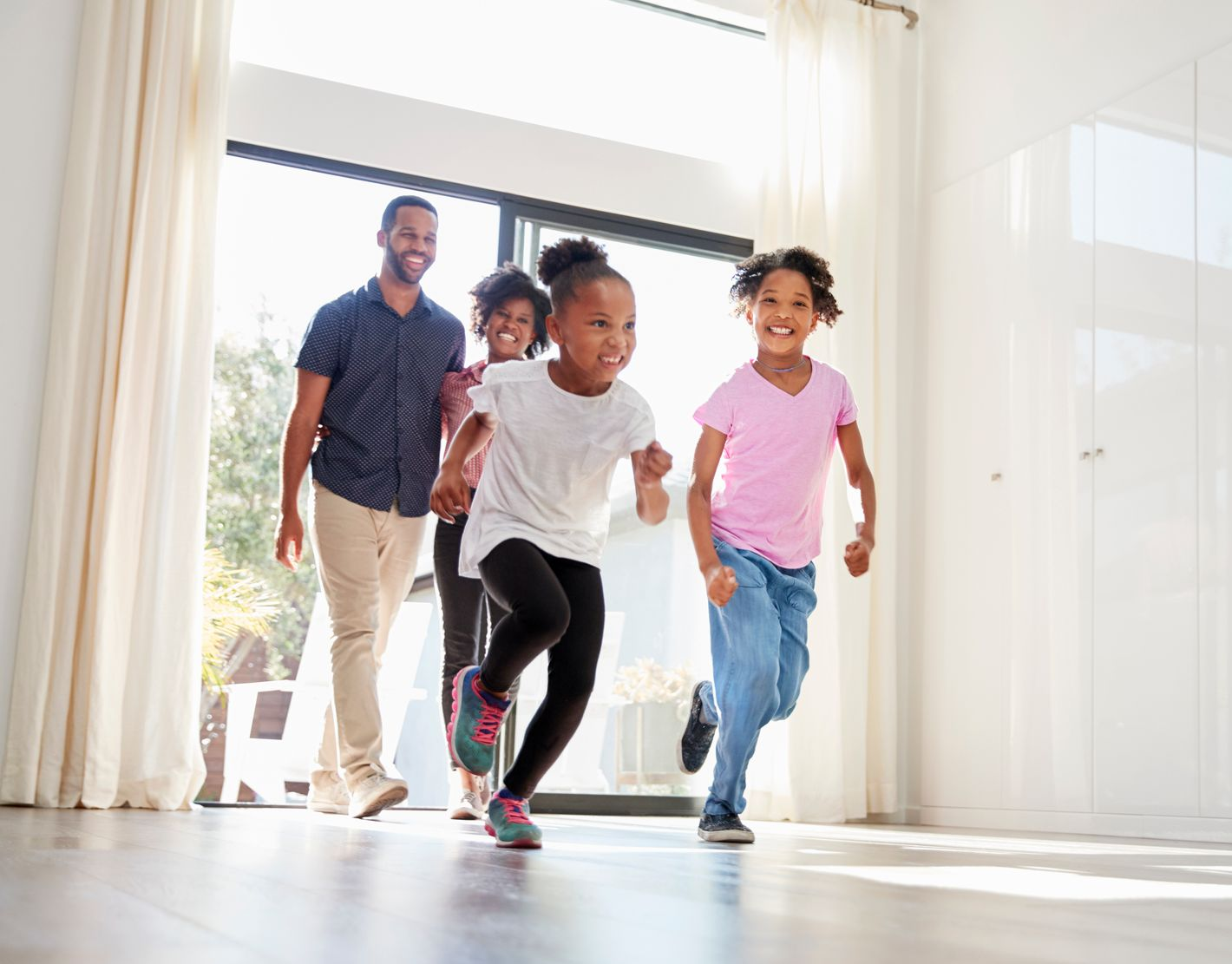 Two children run joyfully into a sunlit room, followed by two adults walking through an open glass doorway.