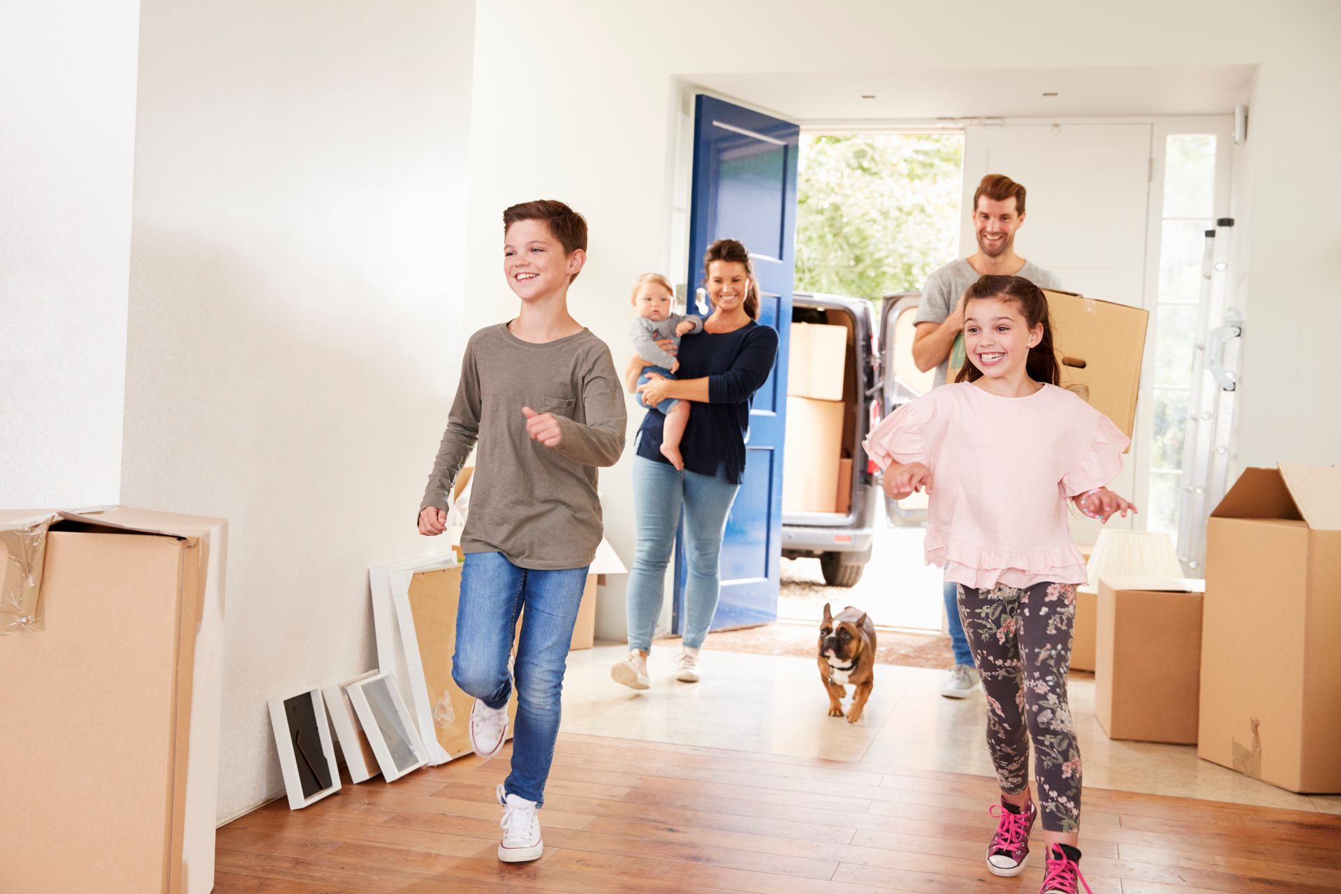 A family with two children and a dog moves into a new house, carrying boxes into an entryway filled with cardboard crates.