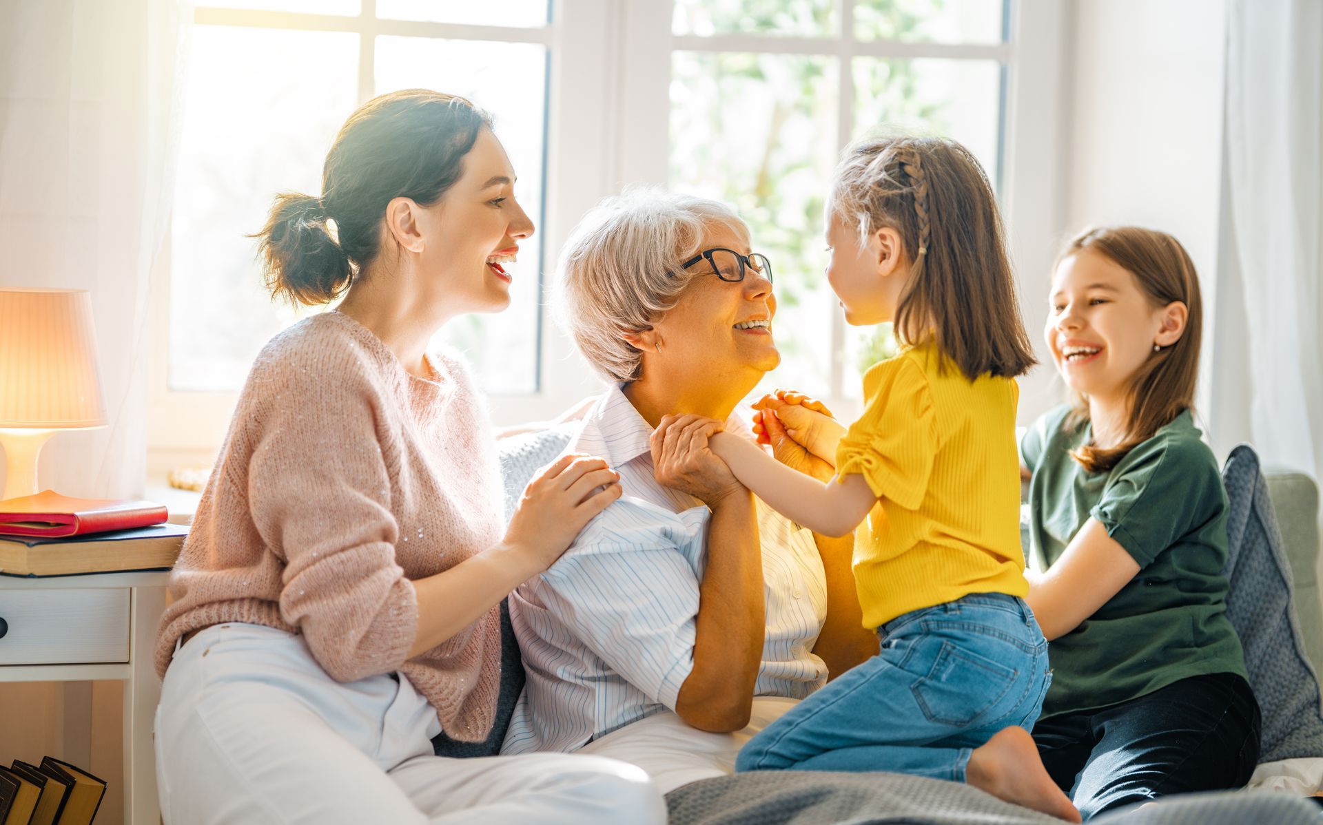 A multi-generational family sitting together indoors by a window, smiling and interacting affectionately.