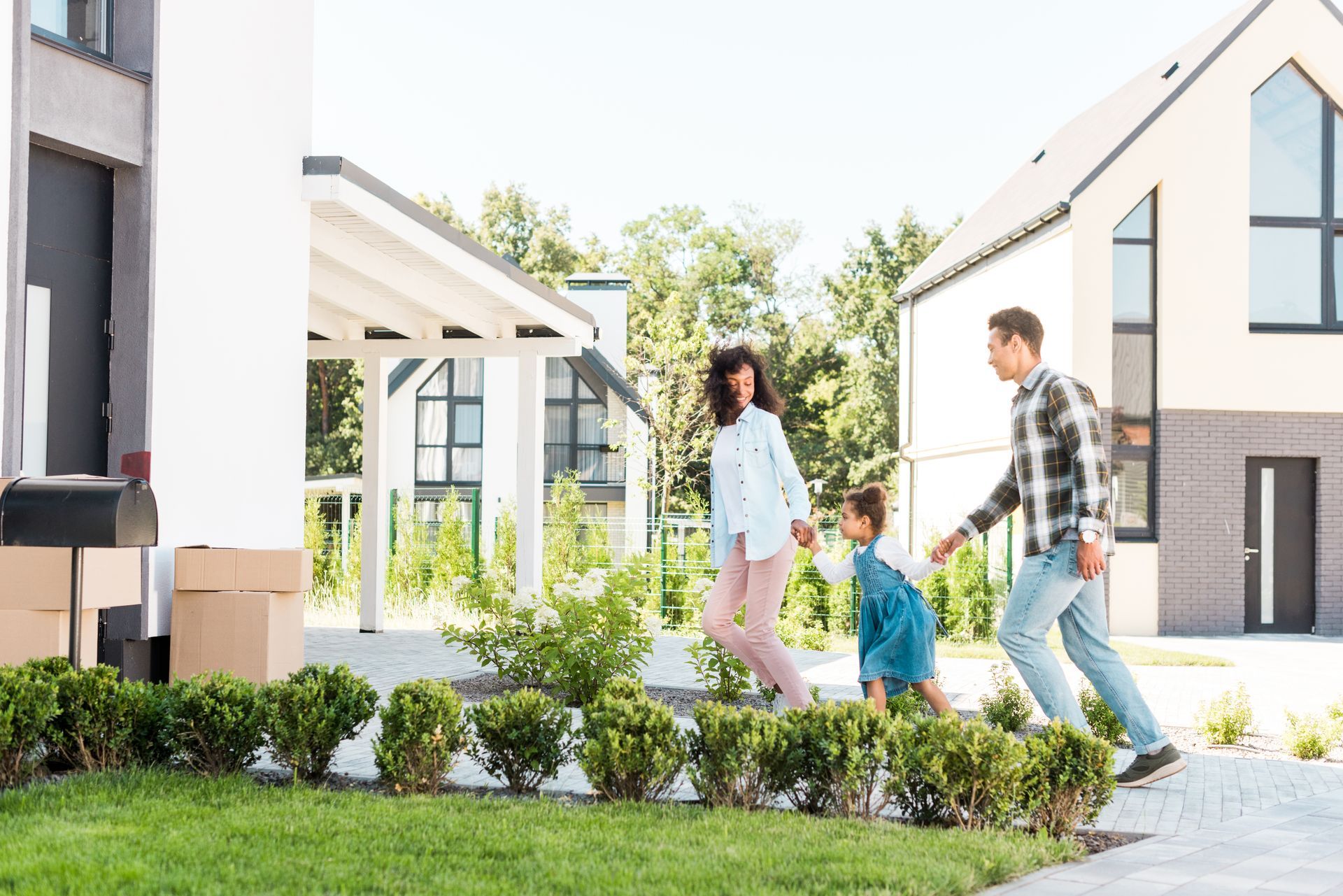 A family of three walking and holding hands along a paved path in a modern residential neighborhood on a sunny day.