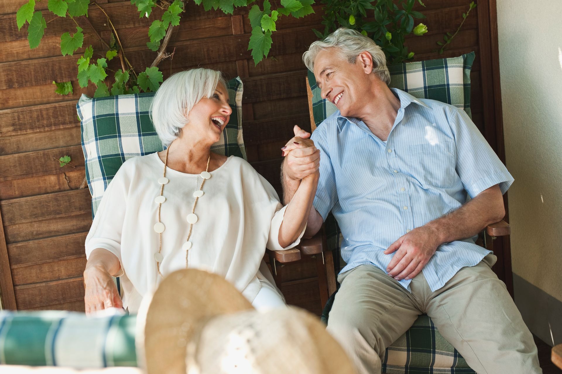 A smiling couple sitting on a wooden bench holding hands, with a green vine in the background.