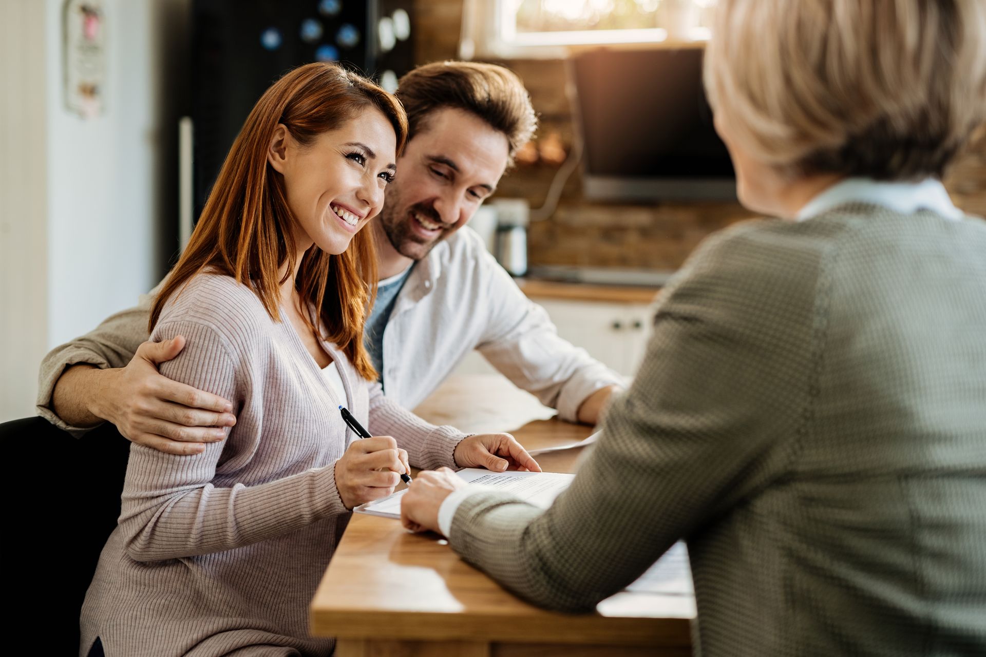 A smiling couple sits at a wooden table reviewing documents with an advisor in a bright, modern office setting.
