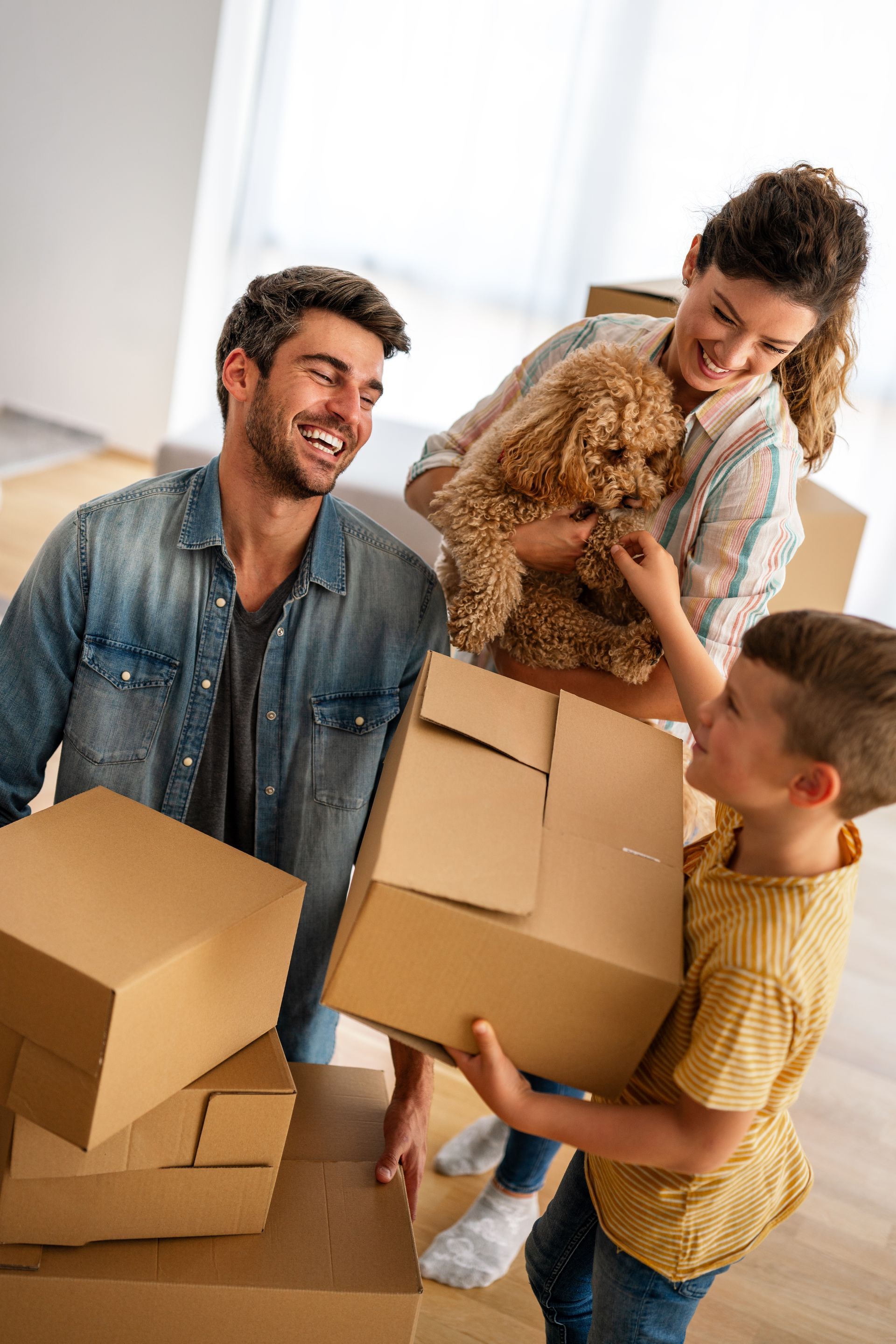 A family with their dog holds cardboard boxes in a bright, unfurnished room while moving into a new home.