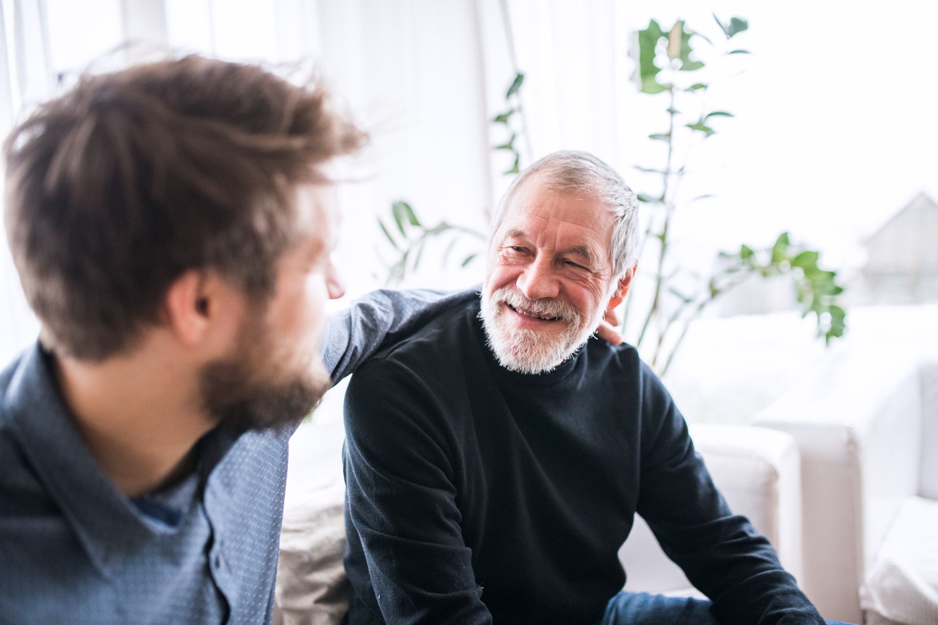 Two people sit together, one with a hand on the other's shoulder, sharing a warm conversation in a bright, indoor space.