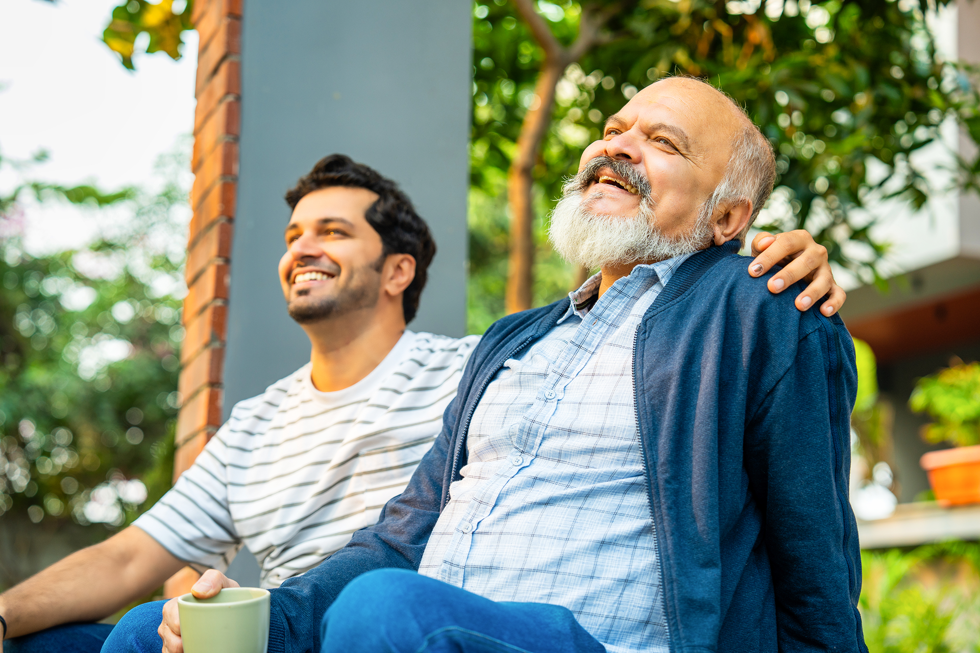 Two men smiling and sitting outdoors, with one man resting his arm on the other’s shoulder in a warm, relaxed moment.