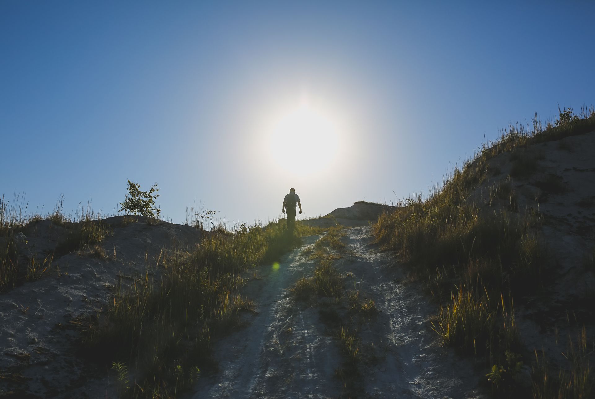 A silhouette of a person hiking up a steep, grassy, and rocky hill toward the bright, shining sun in a clear blue sky.