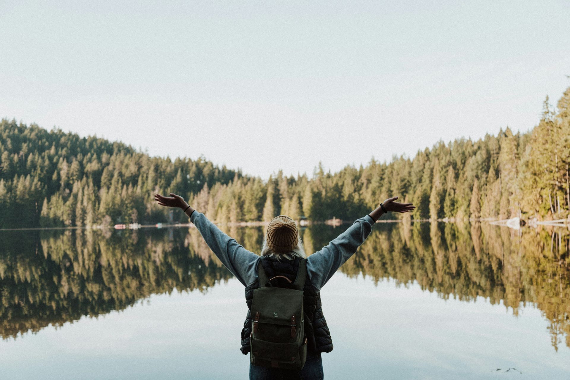 Person with a backpack standing with arms raised before a calm lake reflecting a dense evergreen forest.