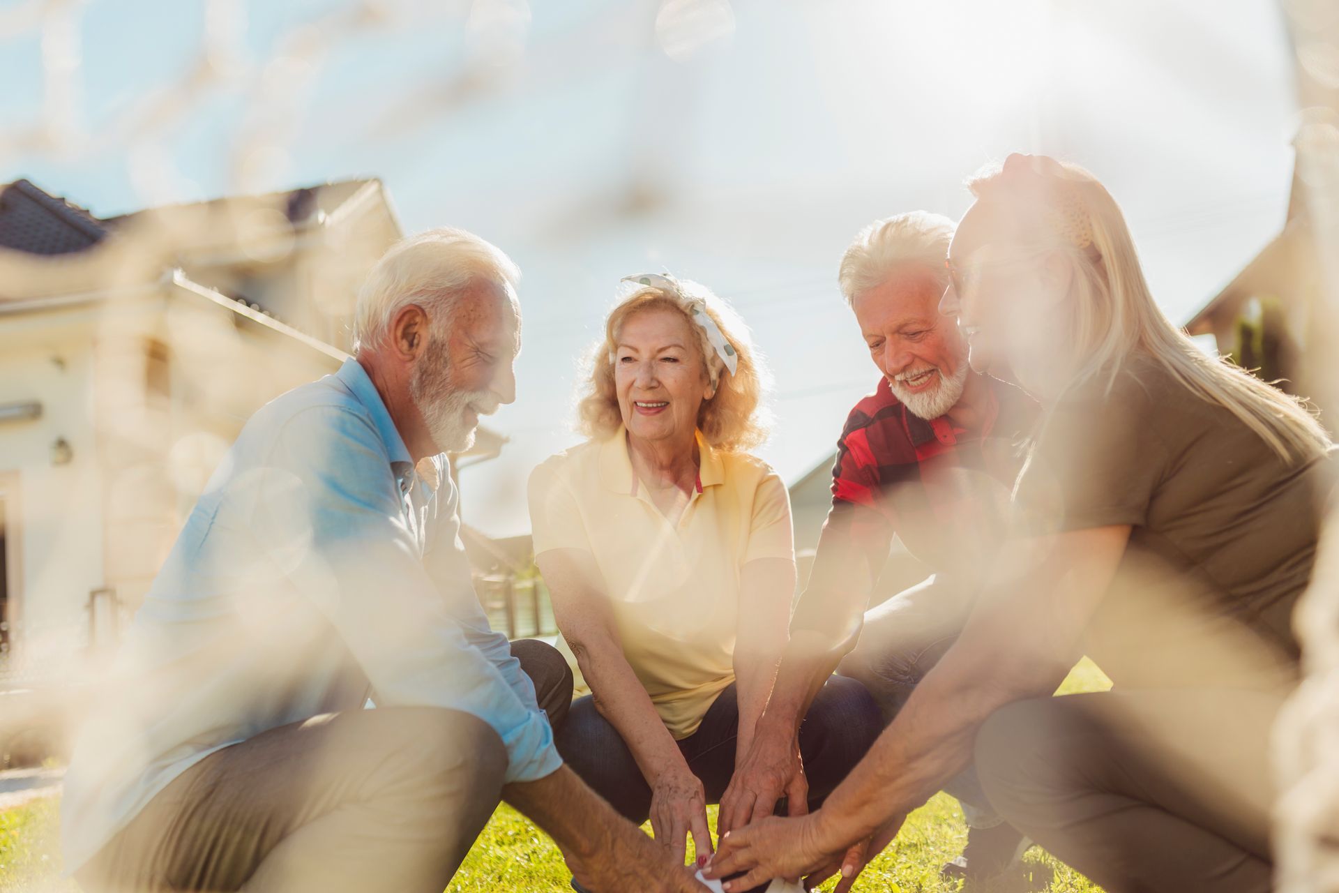 Four people gather in a circle outdoors, joining hands over green grass with a soft, bright lens flare.