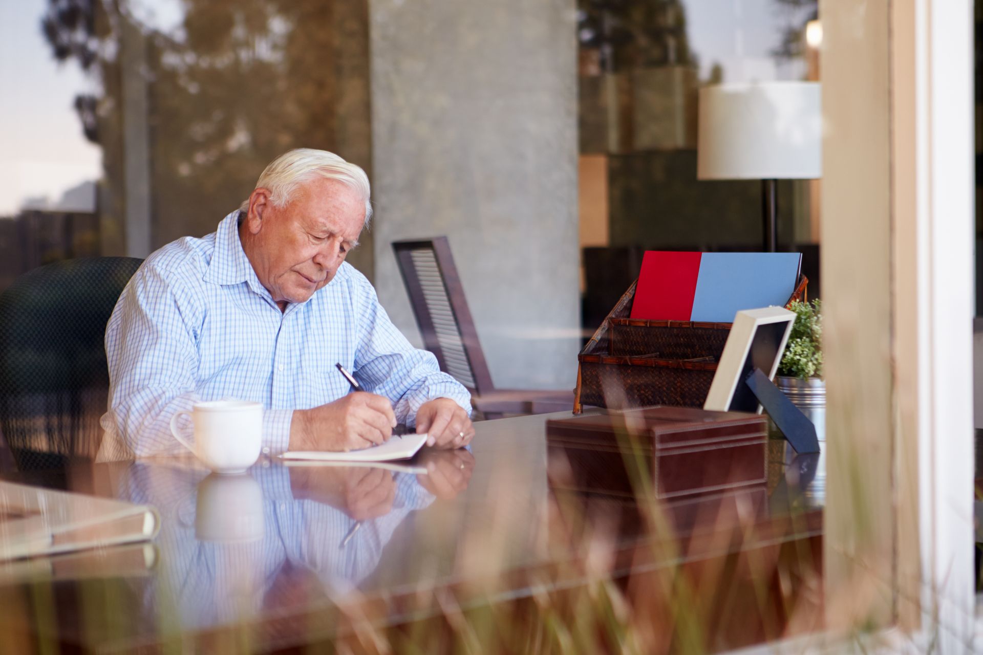 An individual sits at a wooden desk by a window, writing in a notebook next to a white mug.