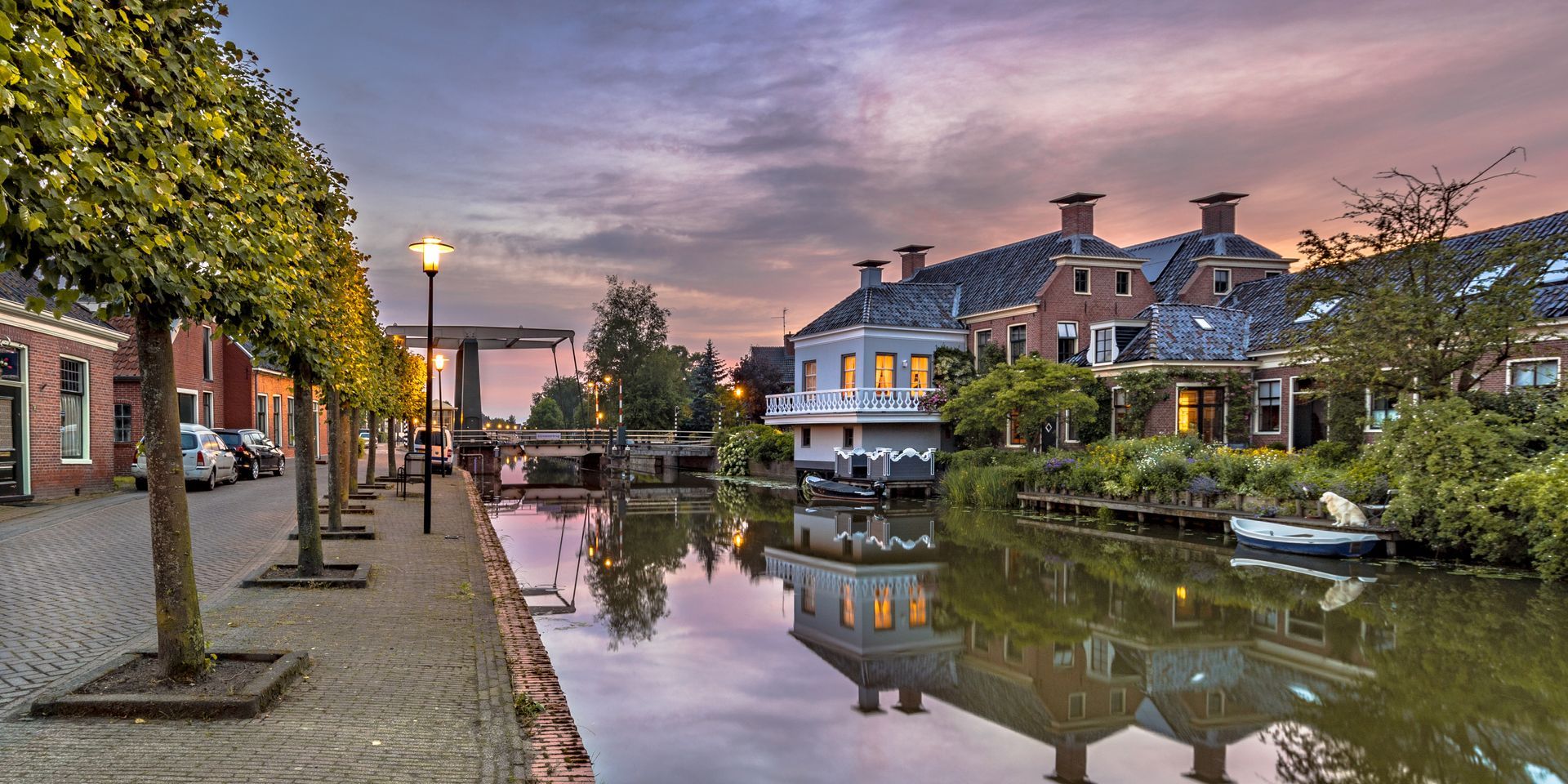 A tree-lined cobblestone street runs beside a canal reflecting historic brick houses under a purple sunset sky.