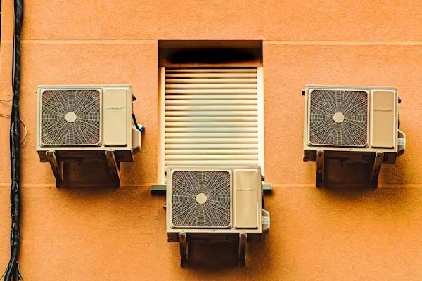 Three HVACs mounted on an orange wall around a shuttered window.