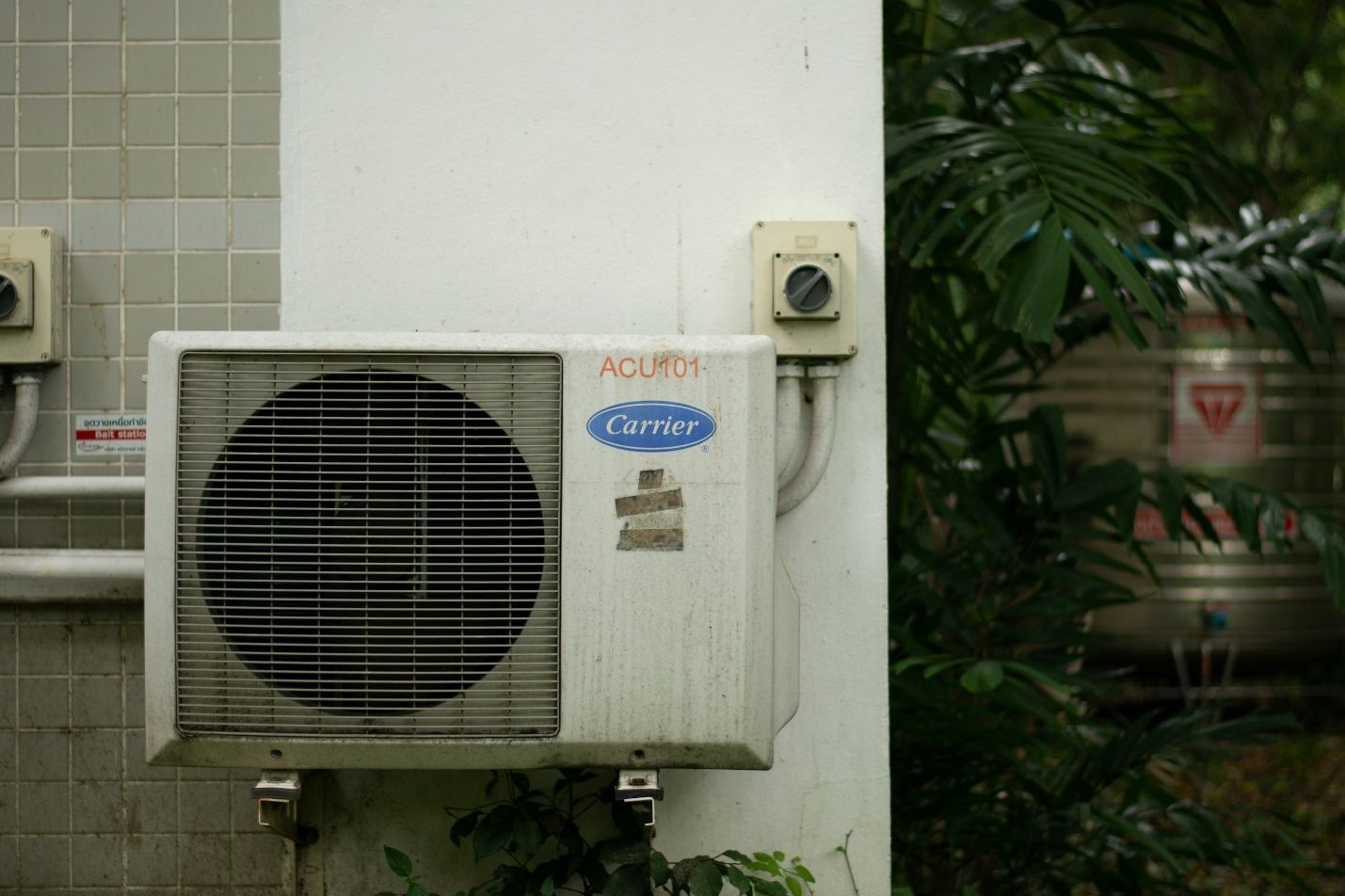 An HVAC outdoor system mounted on a wall with a plant in the background.