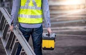 a worker in a yellow vest is carrying a toolbox and a ladder.