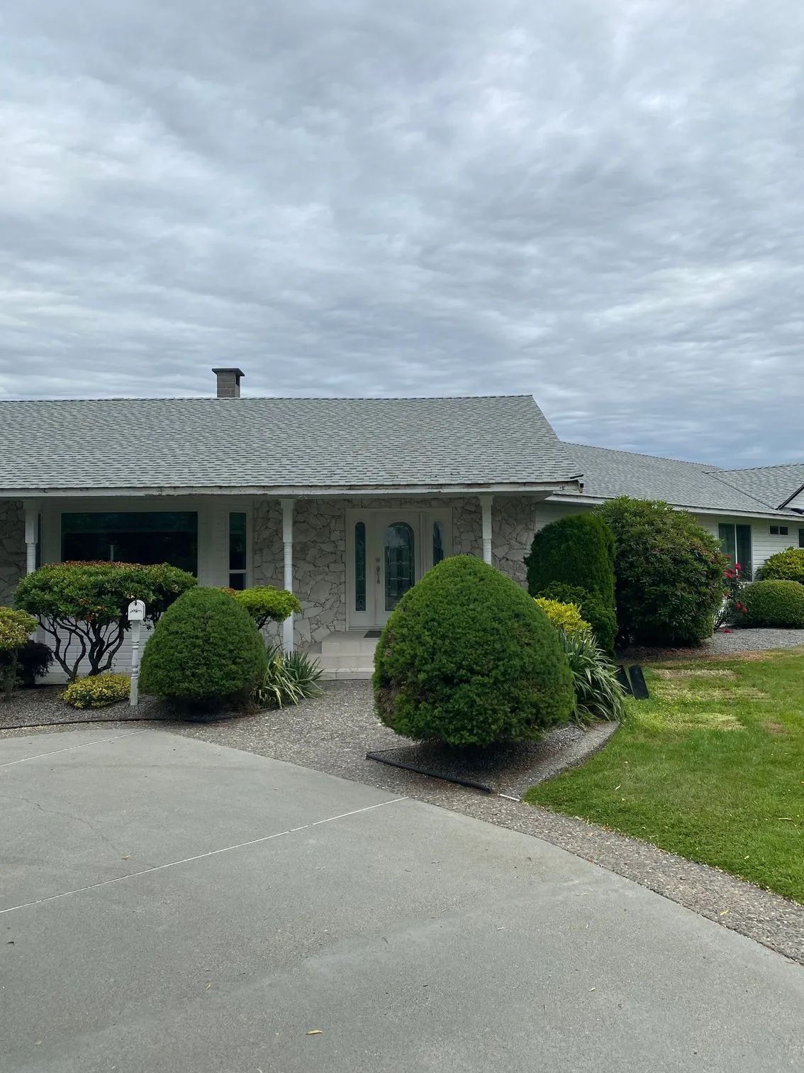 Single-story suburban house with a curved driveway, trimmed shrubs, and a cloudy sky.