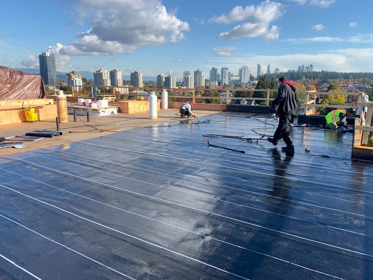 Workers installing roofing membrane on a rooftop with a city skyline in the background.