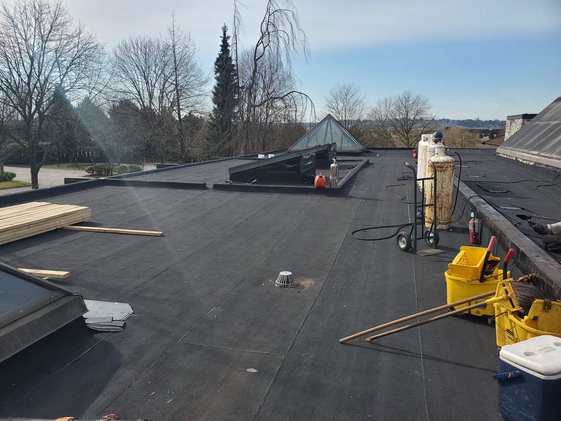 Flat rooftop with roofing materials, tools, and workers repairing the surface under a clear sky