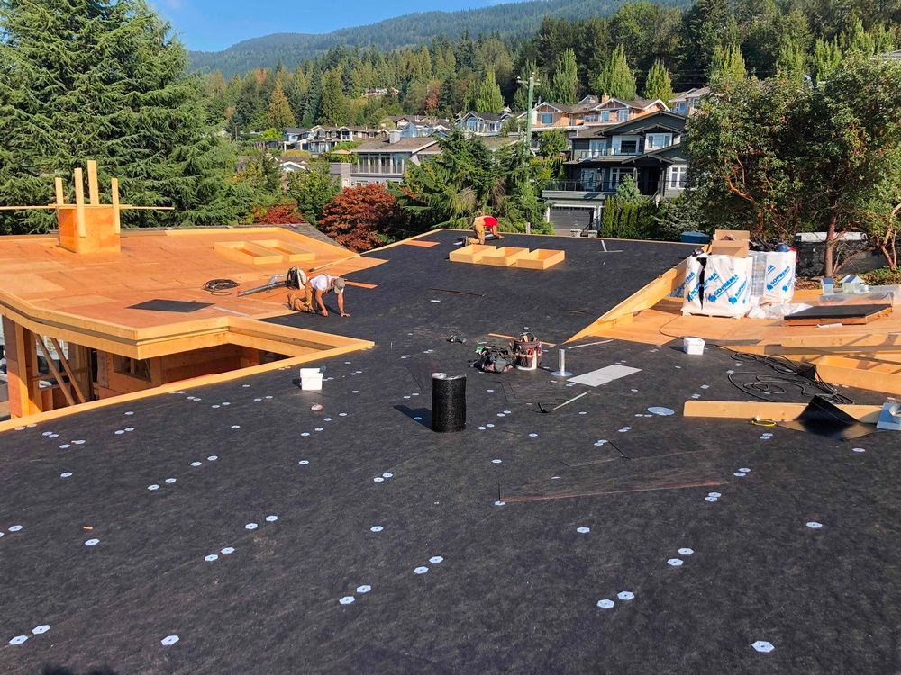 Construction workers on a roof install black roofing material on a sunny day. Houses and trees in the background.