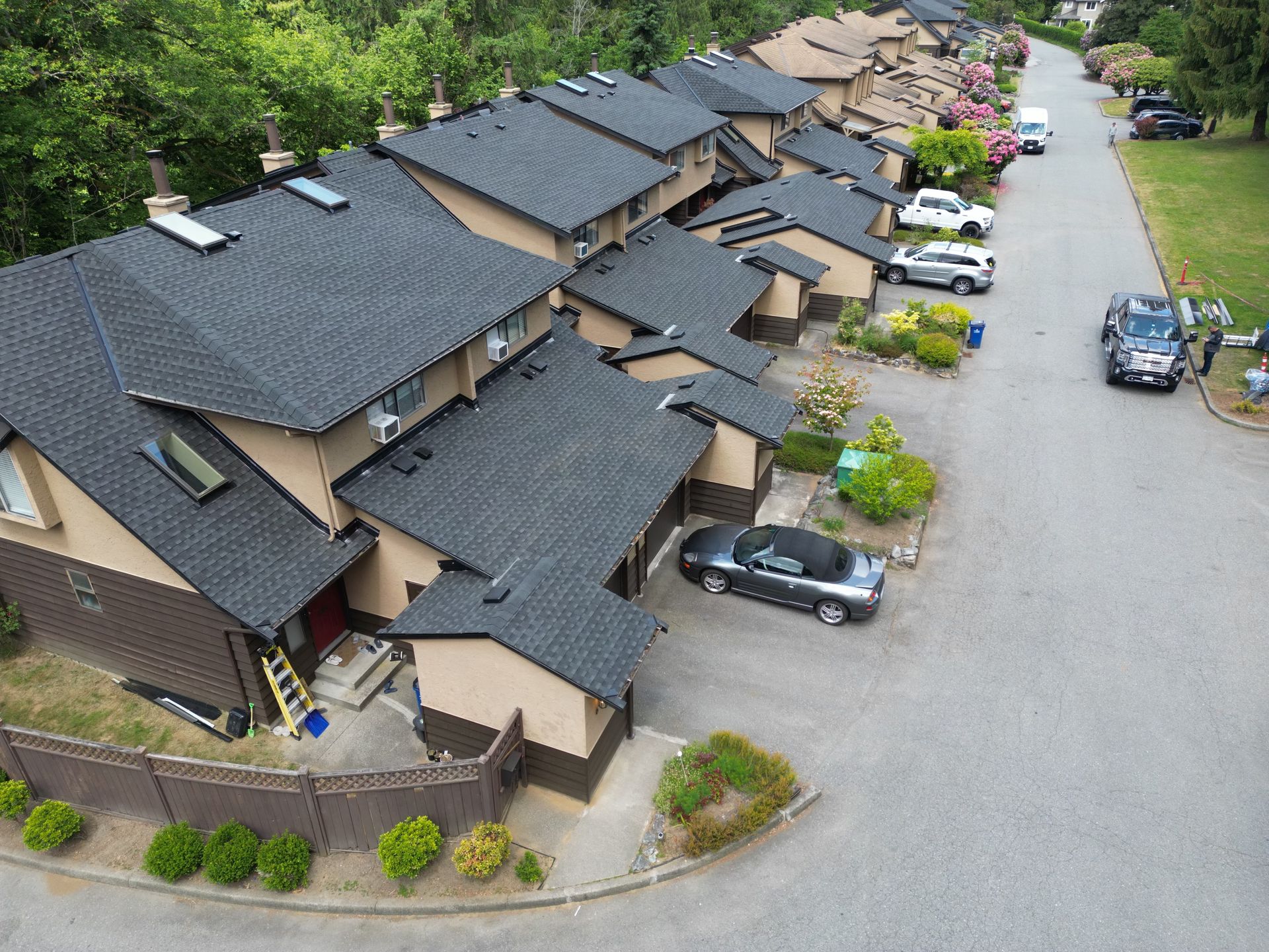 Row of townhouses with dark roofs and parked cars along a street, sunny day.