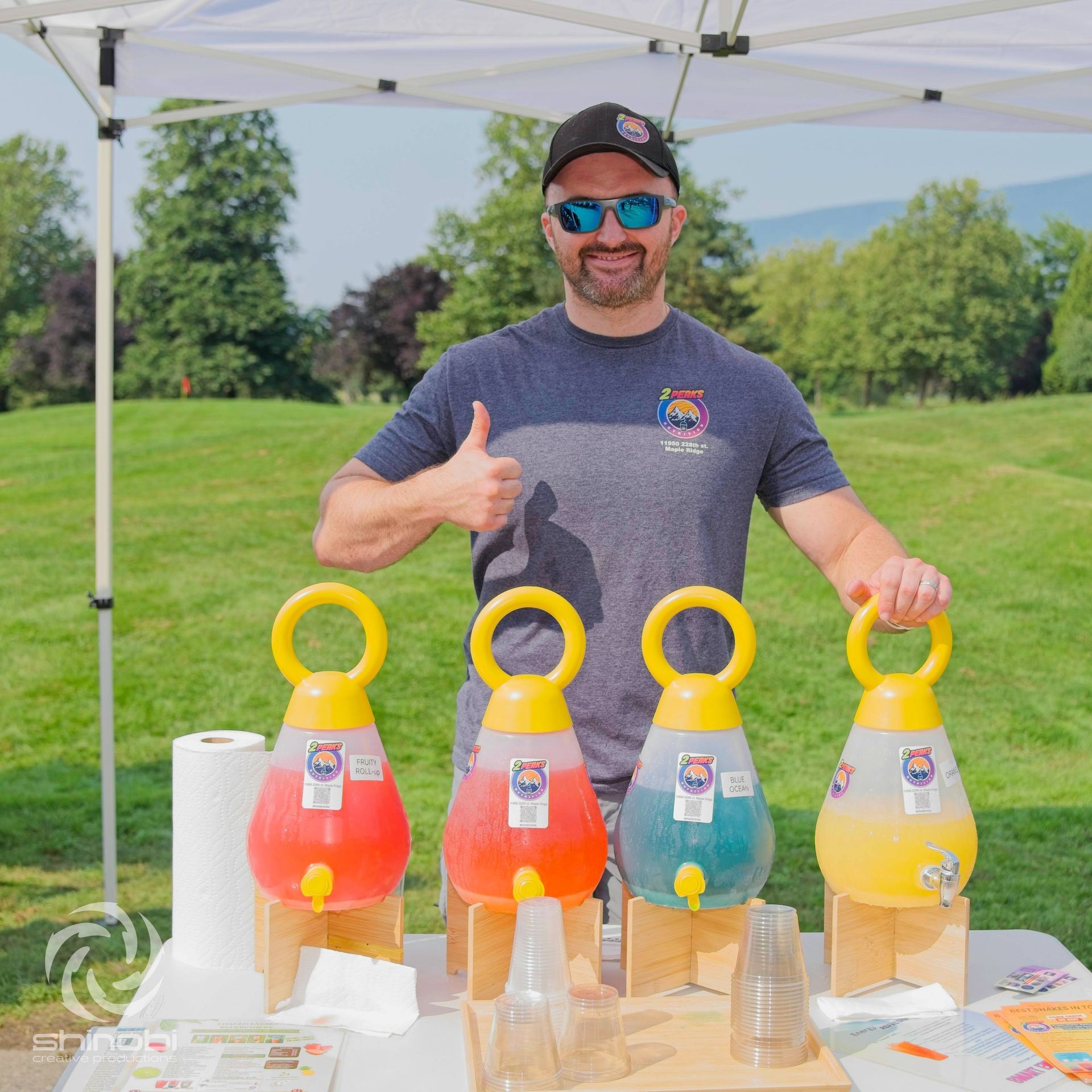 A man with sunglasses and a thumbs up stands behind a table with four colorful drink dispensers.