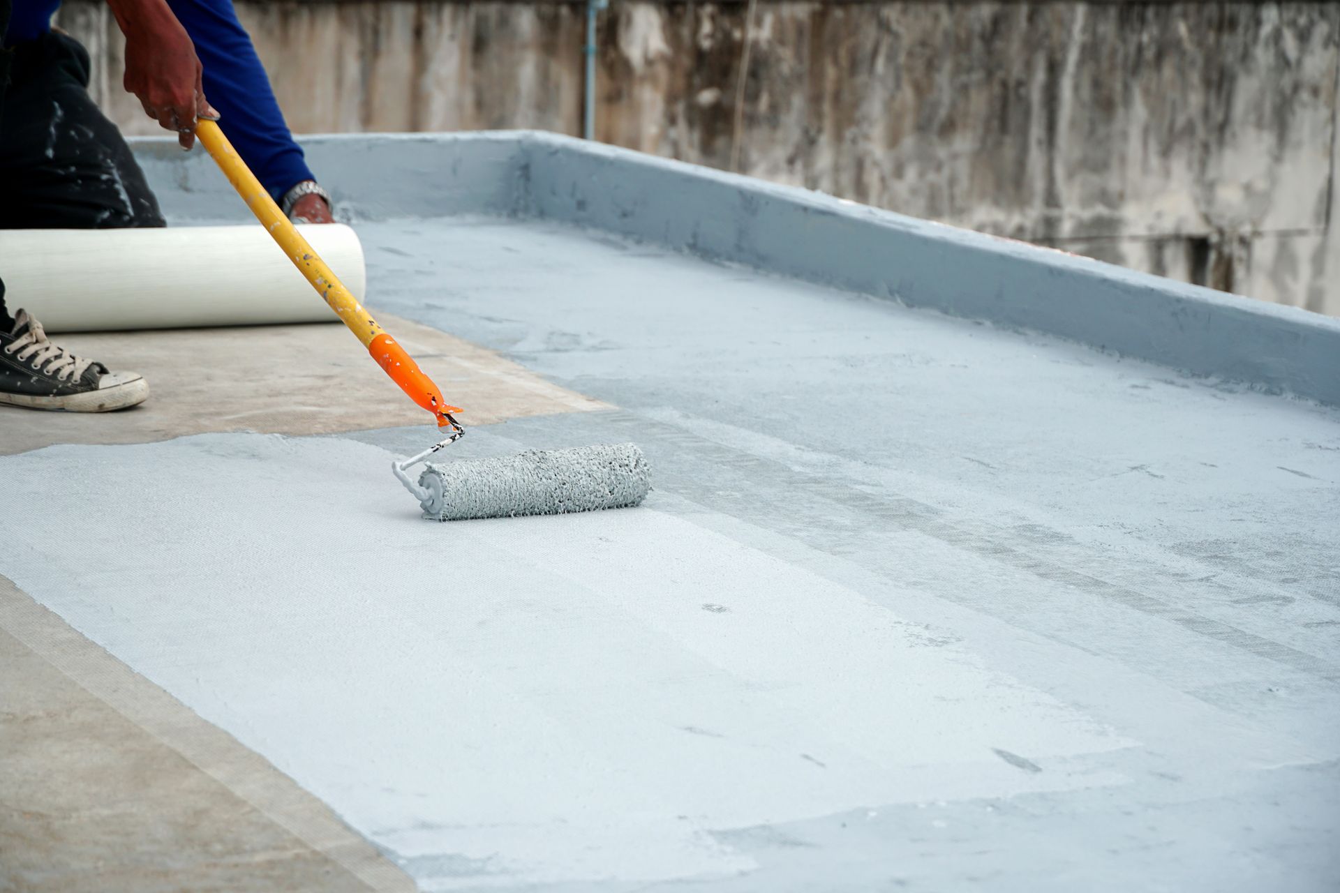 A person applying a grey sealant to a flat roof with a roller. A roll of material sits nearby.