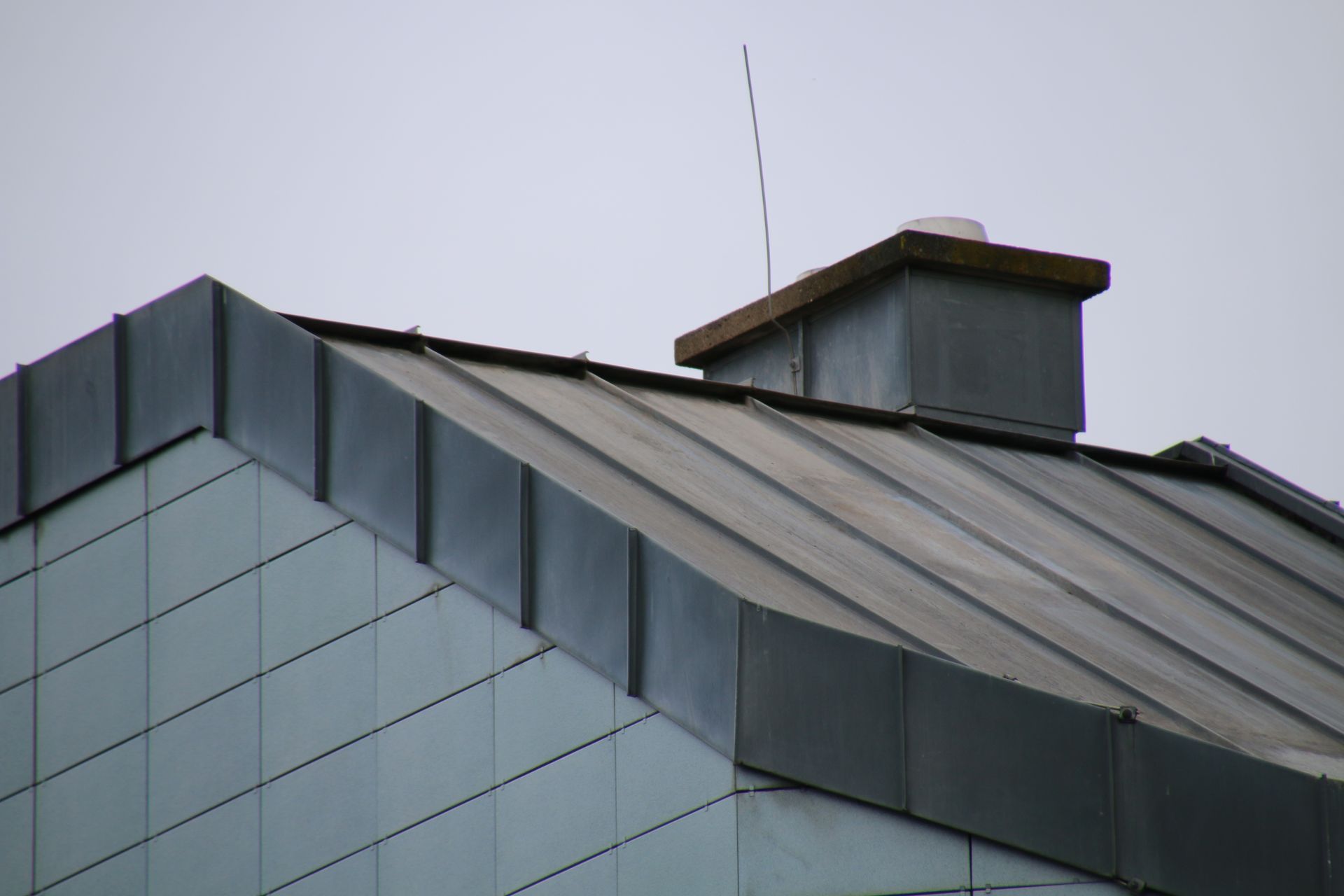 A dark gray metal roof with a chimney and lightning rod on a cloudy day.
