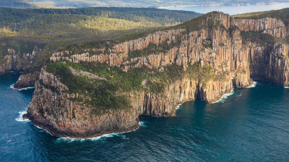 Cliffside landscape with towering rock formations meeting the blue ocean. Green foliage atop cliffs, sunny day.