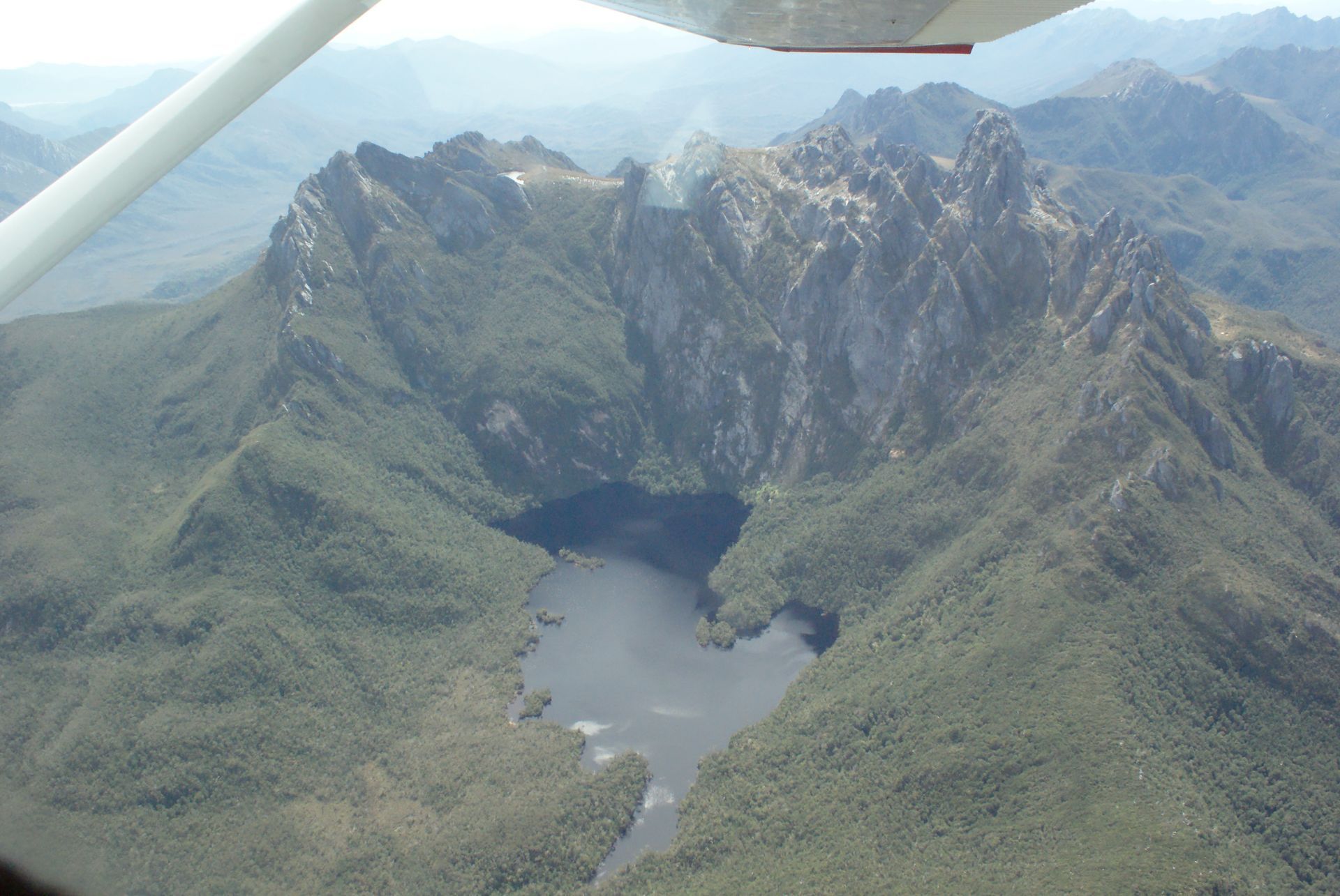 Aerial view: jagged mountain peaks with a dark lake surrounded by dense green forest.